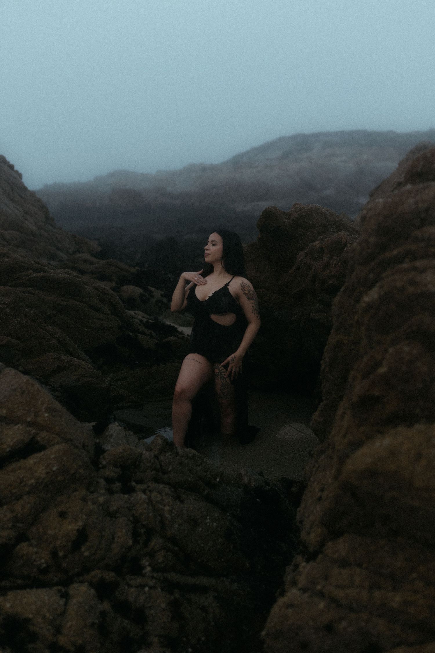 Dark figure perches on rugged coastal rocks at twilight.