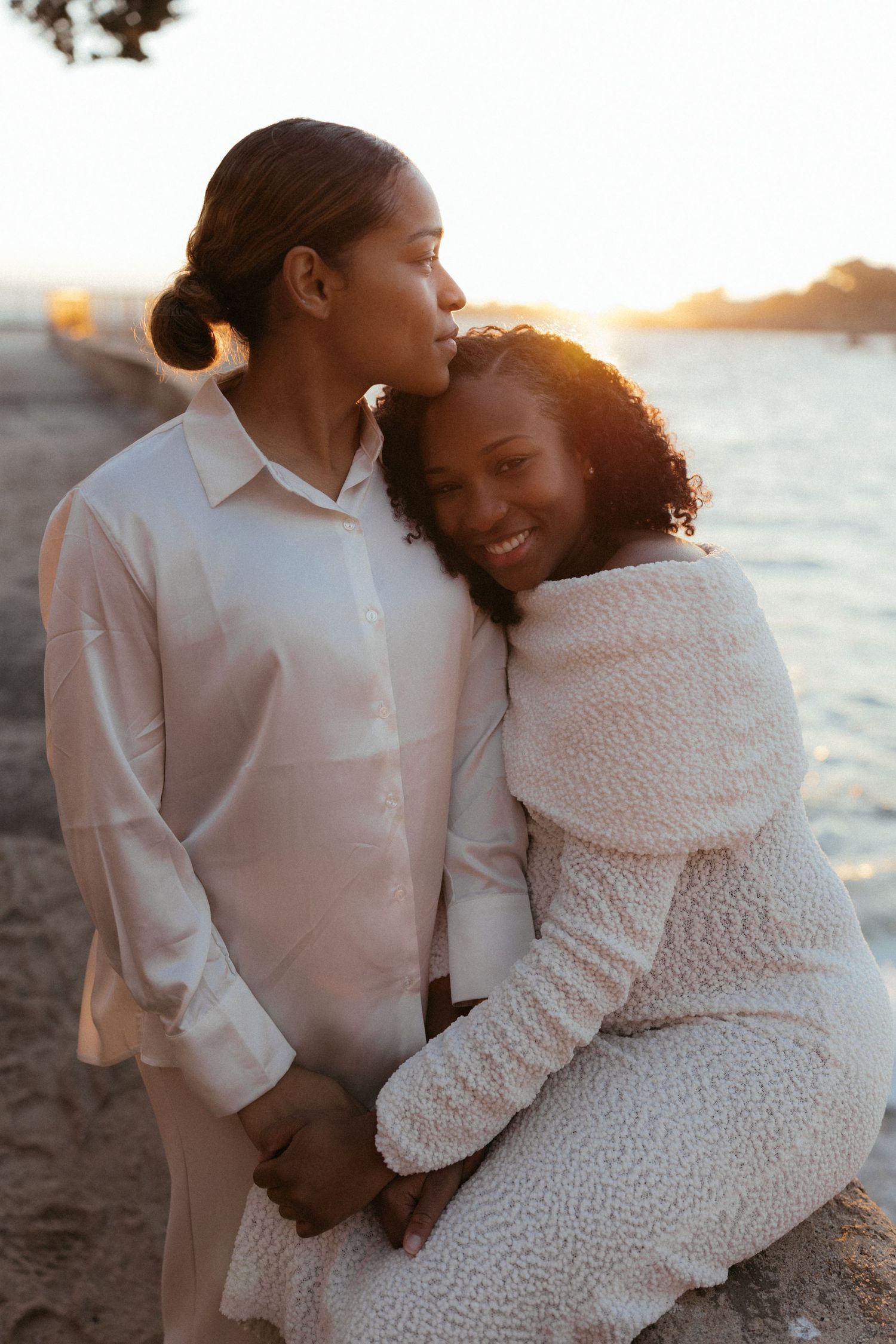 Romantic couple embraces on beach at sunset in white outfits during golden hour photo session.
