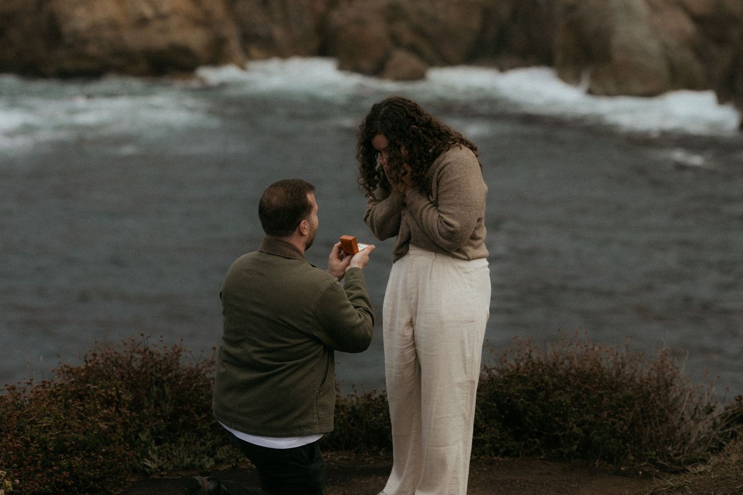 A couple stands near rocky cliffs by the ocean during an engagement photoshoot at sunset.