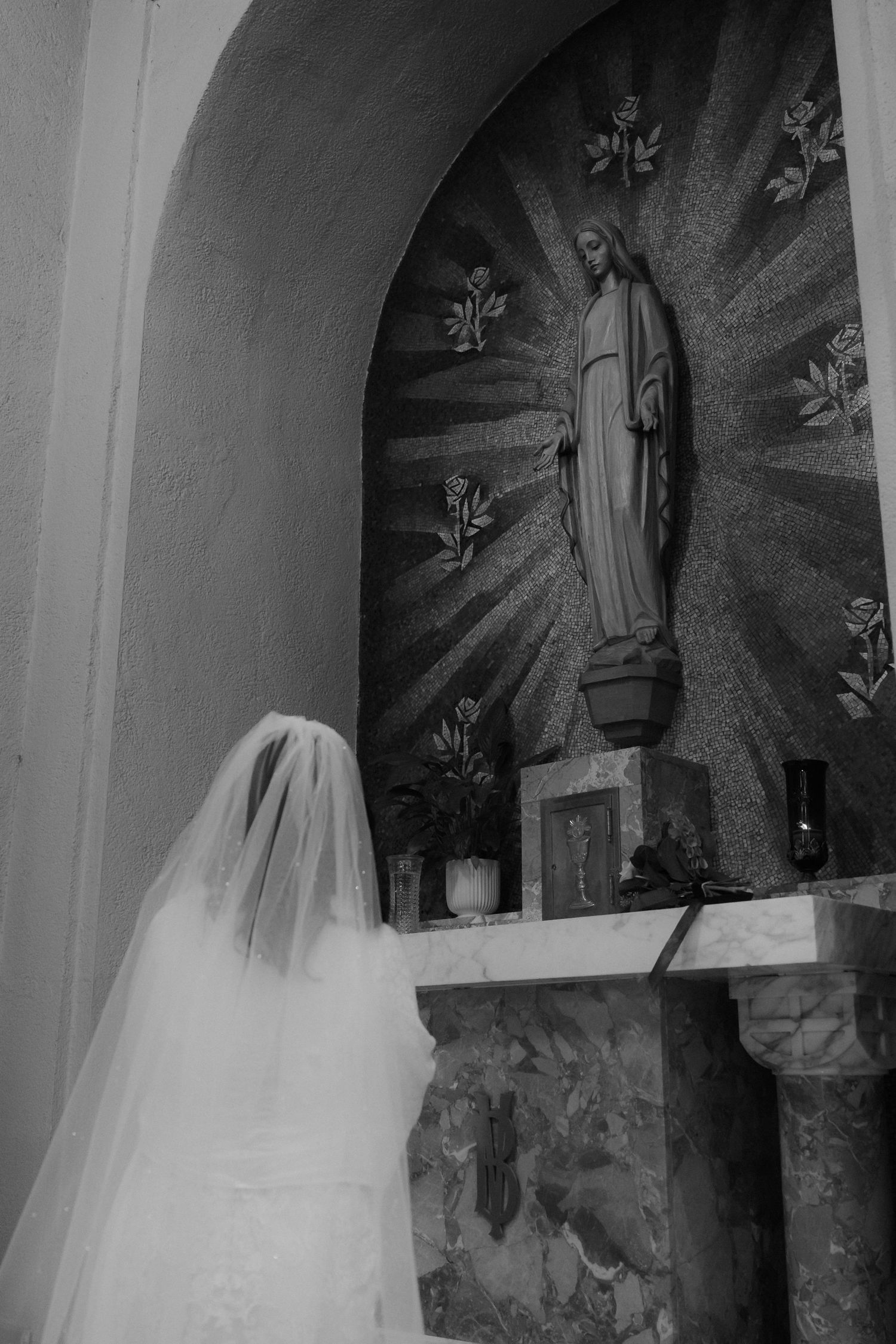Black and white photo of a bride in a flowing veil against church wall art.