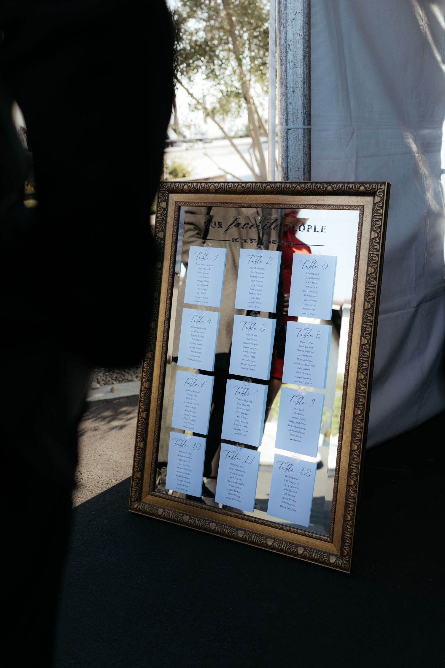 Gold-framed mirror displaying wedding seating arrangements on white paper against a dark backdrop.