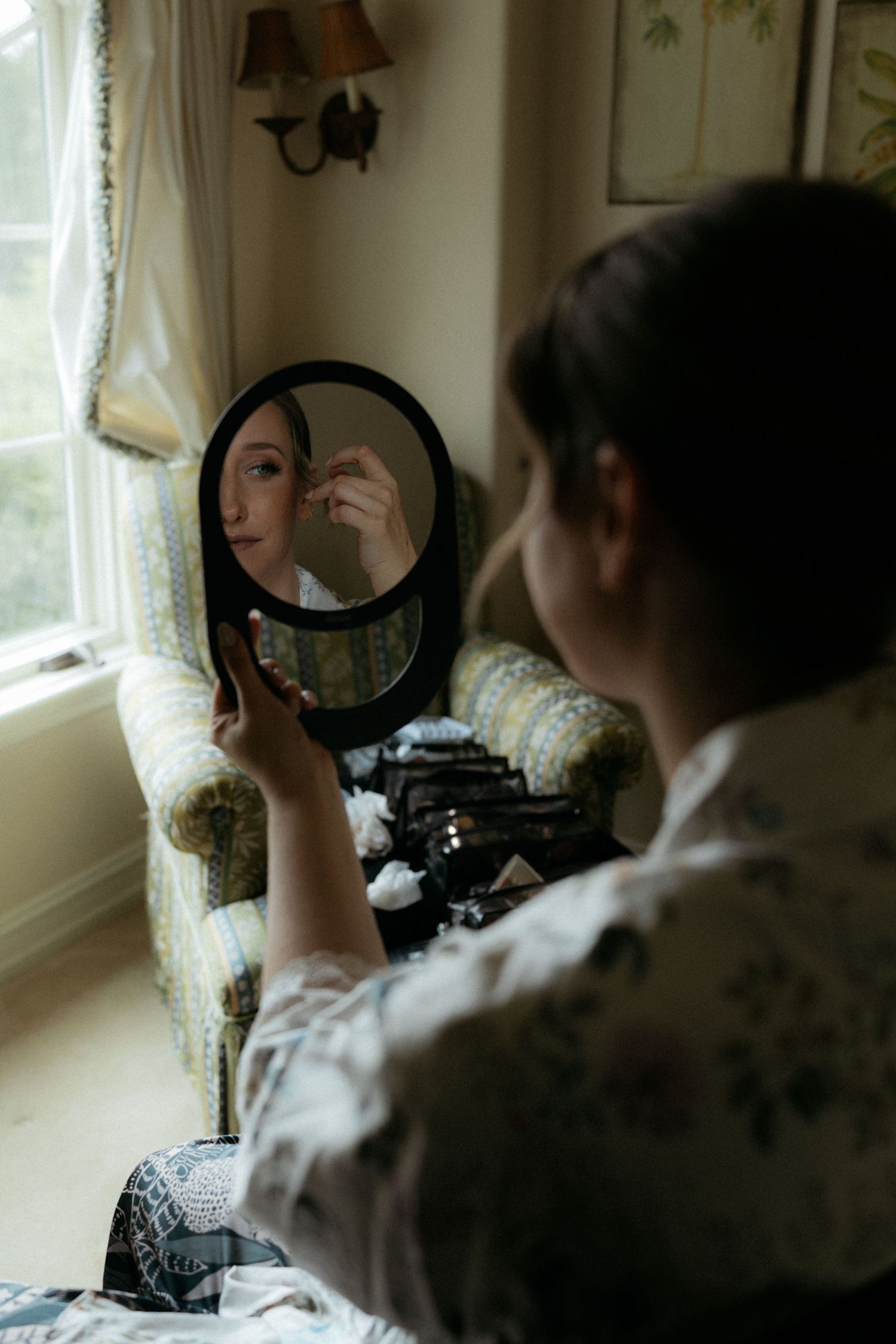 Woman in floral robe applying makeup while holding a round mirror, seated near a bright window in a cozy room.