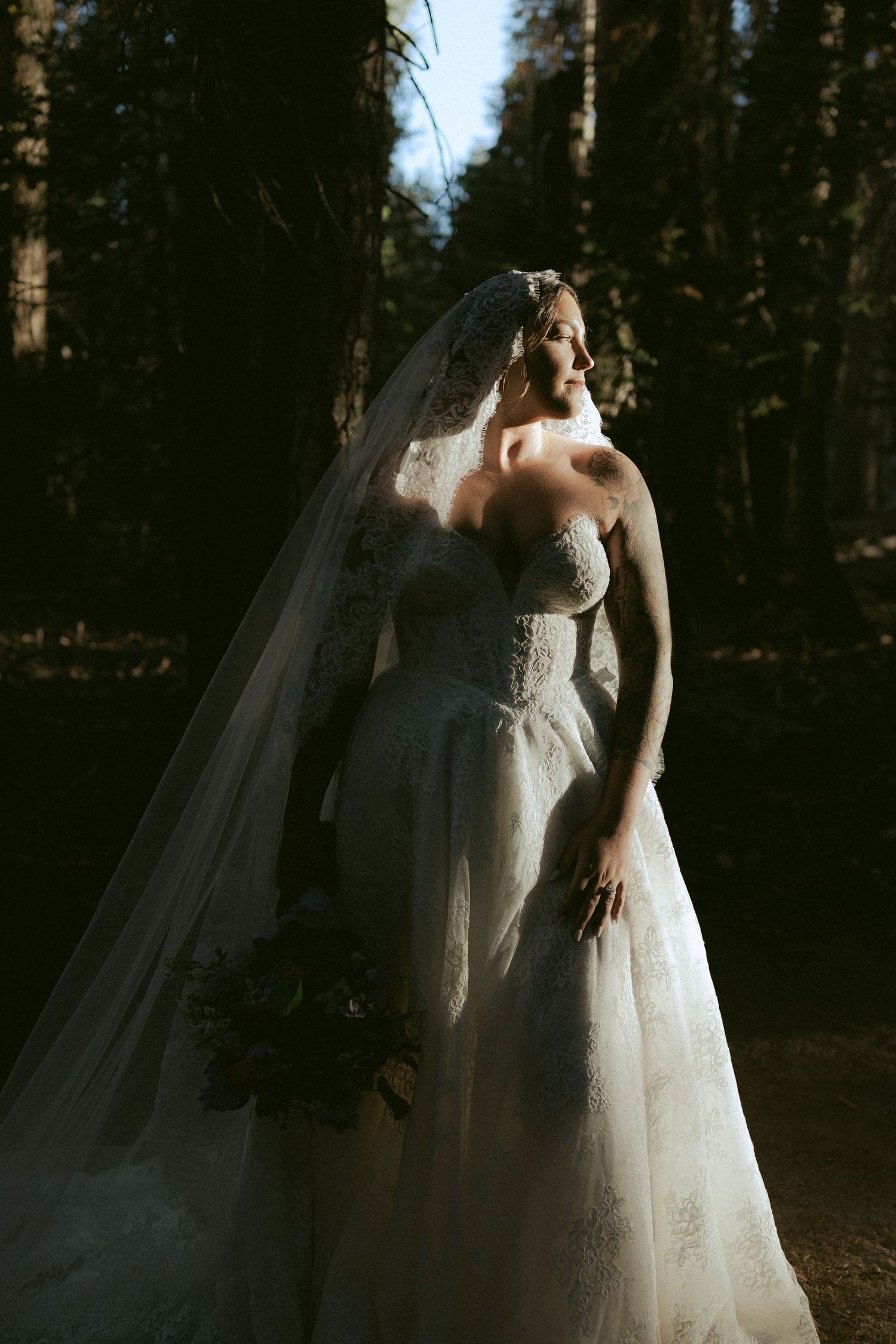Bride in strapless lace wedding gown and veil holding bouquet in sunlit forest.