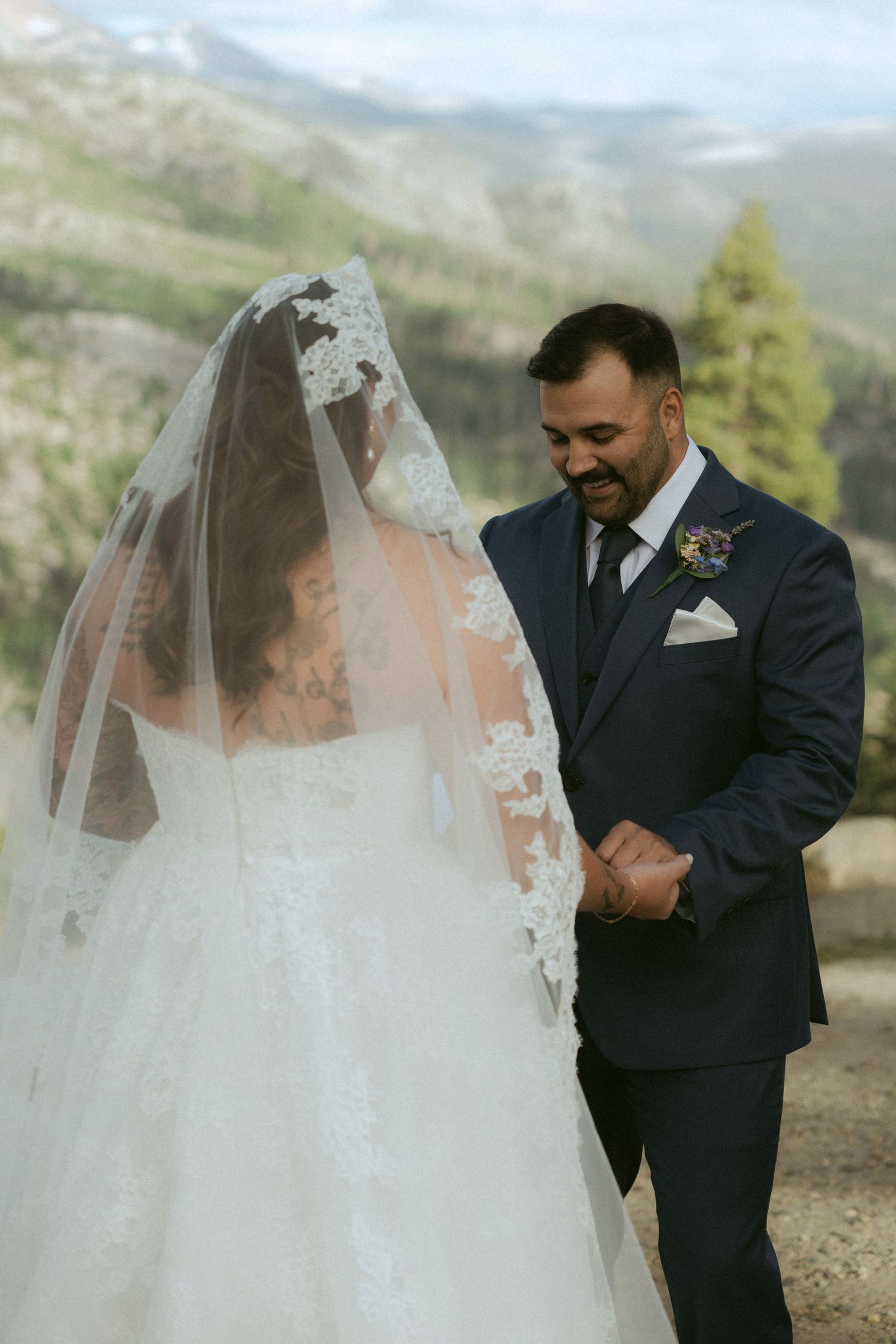 Groom in navy suit smiling at veiled bride during outdoor mountain wedding ceremony.