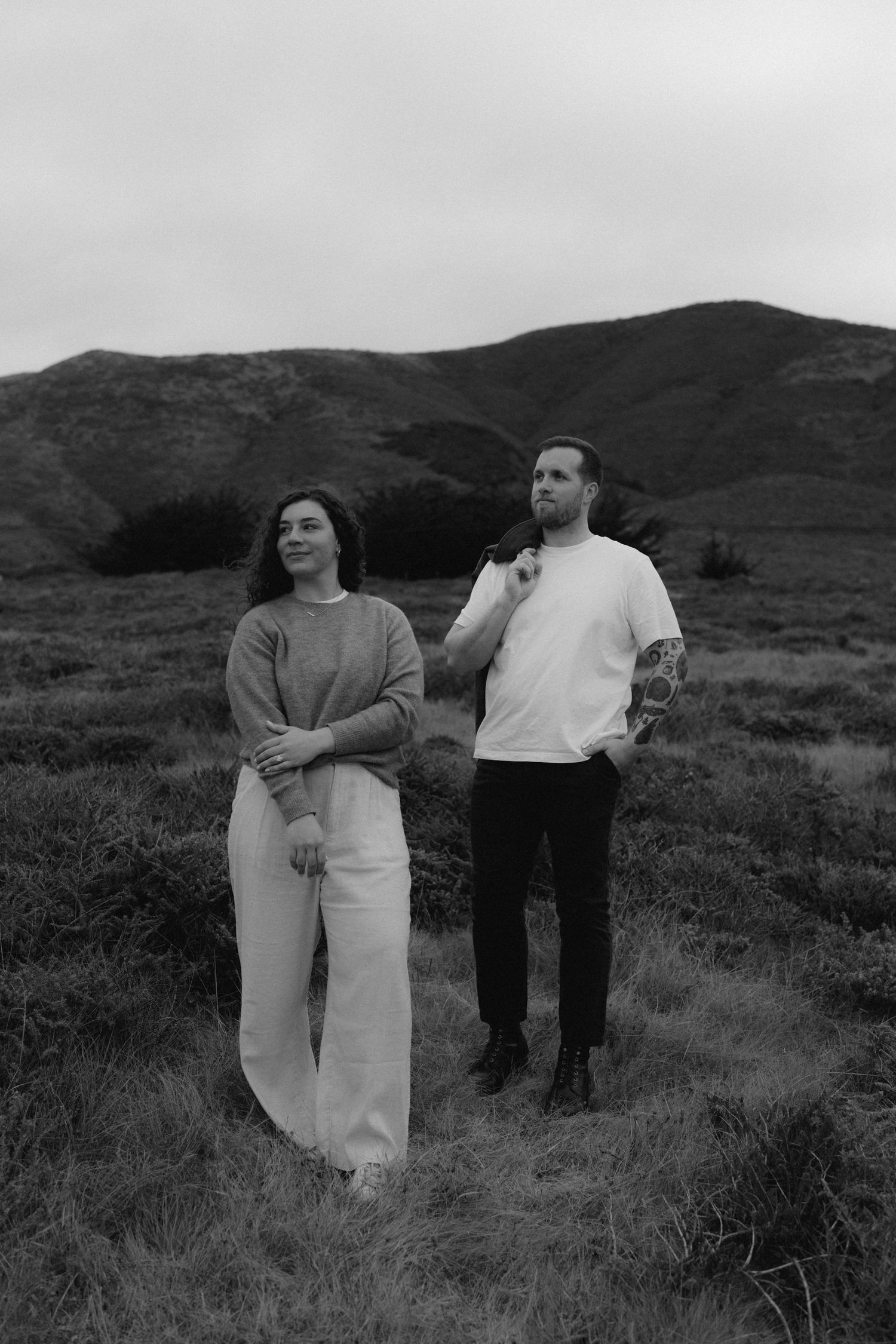 A black and white photo of two people standing together in casual clothing against a mountainous landscape background.