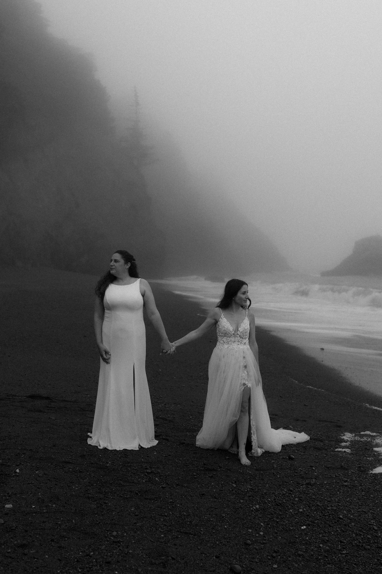 Two figures in white gowns hold hands while walking along a dramatic foggy coastline.