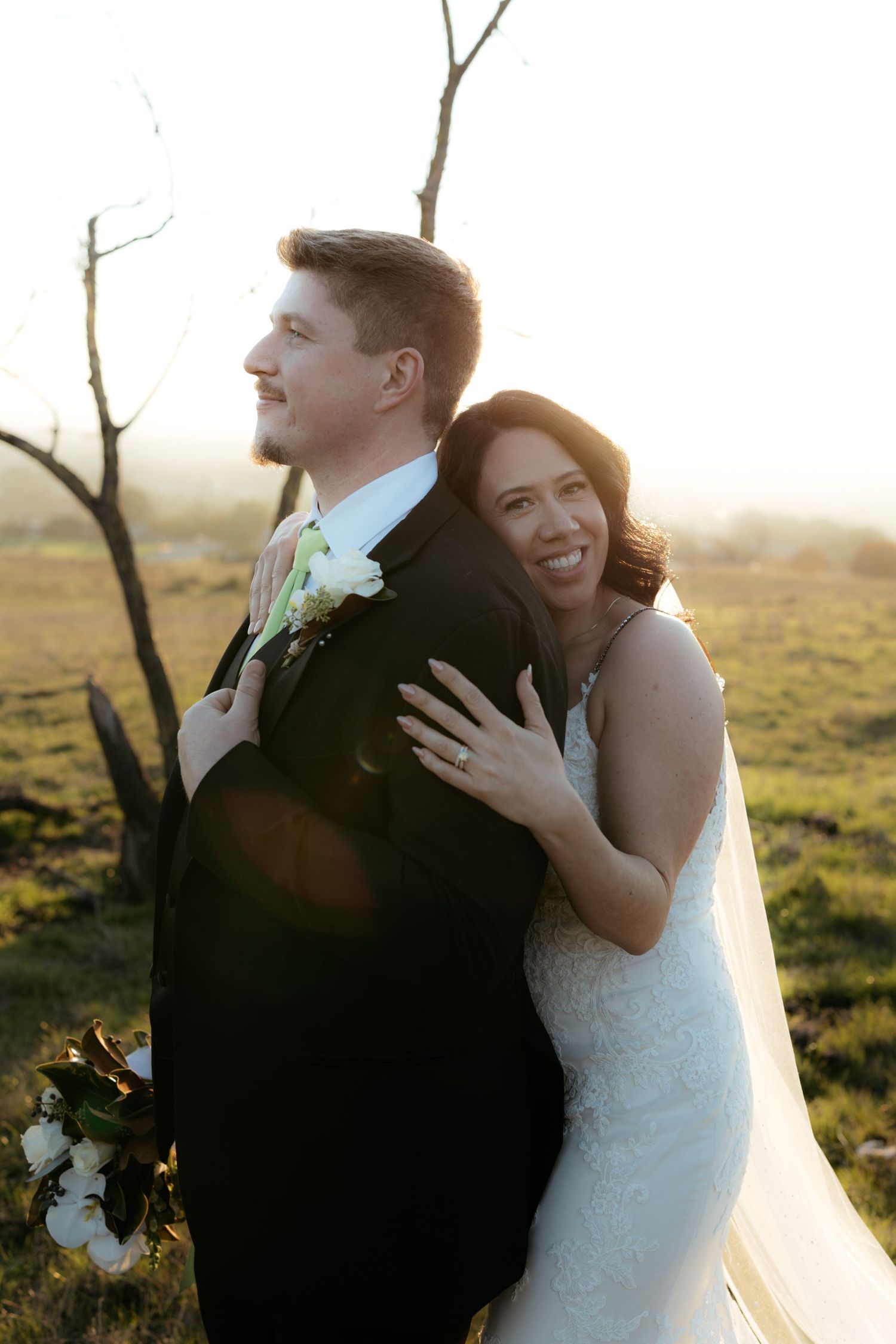 Newlywed couple embracing in golden sunlight with bride in white dress and groom in dark suit.