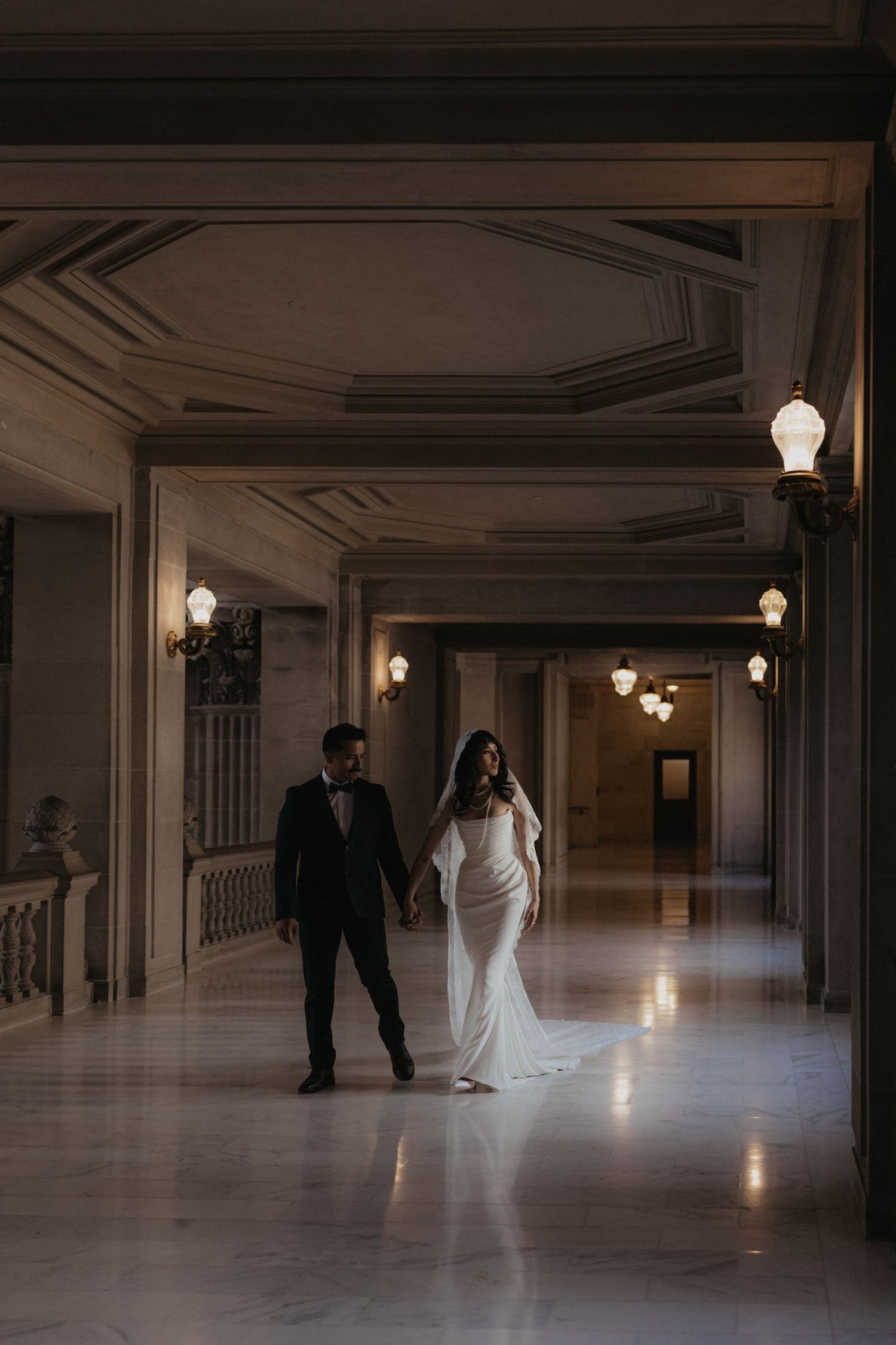 A bride and groom walking through an elegant hallway with ornate ceilings and vintage lighting.