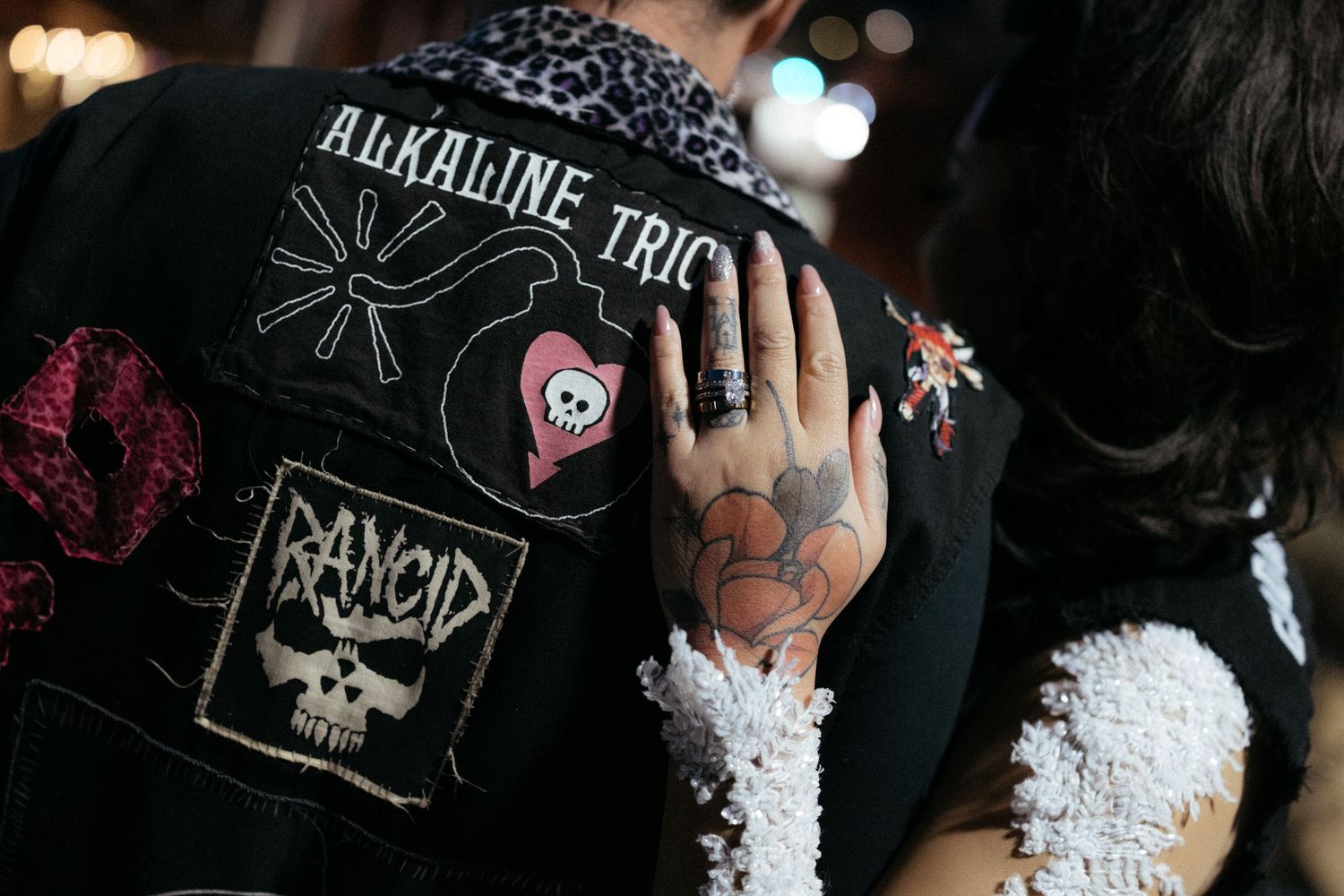 A close-up detail of a punk rock style leather jacket with band patches and white lace accents.