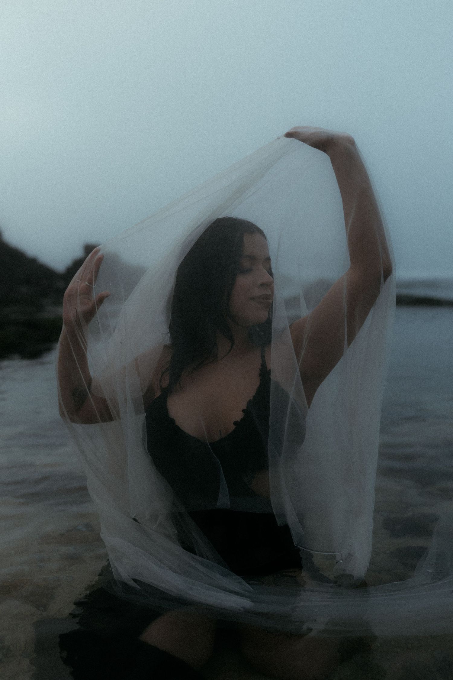 Figure draped in flowing white veil moving gracefully by the water in misty conditions.