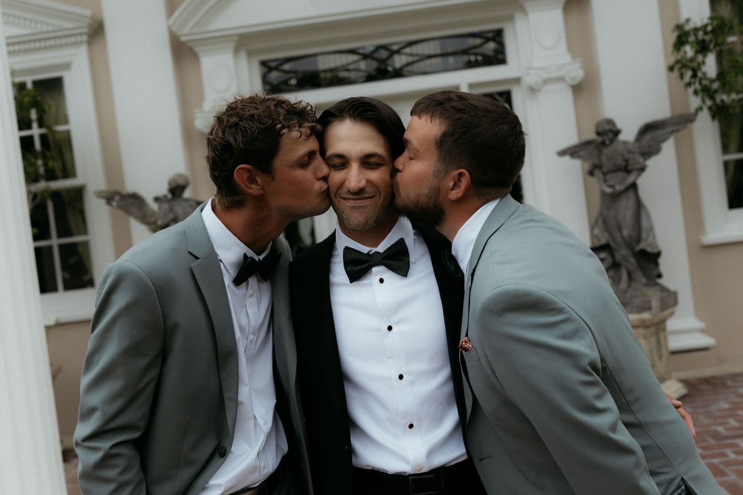 Groom in black tuxedo gets kissed on cheeks by two smiling groomsmen in gray suits outside venue.