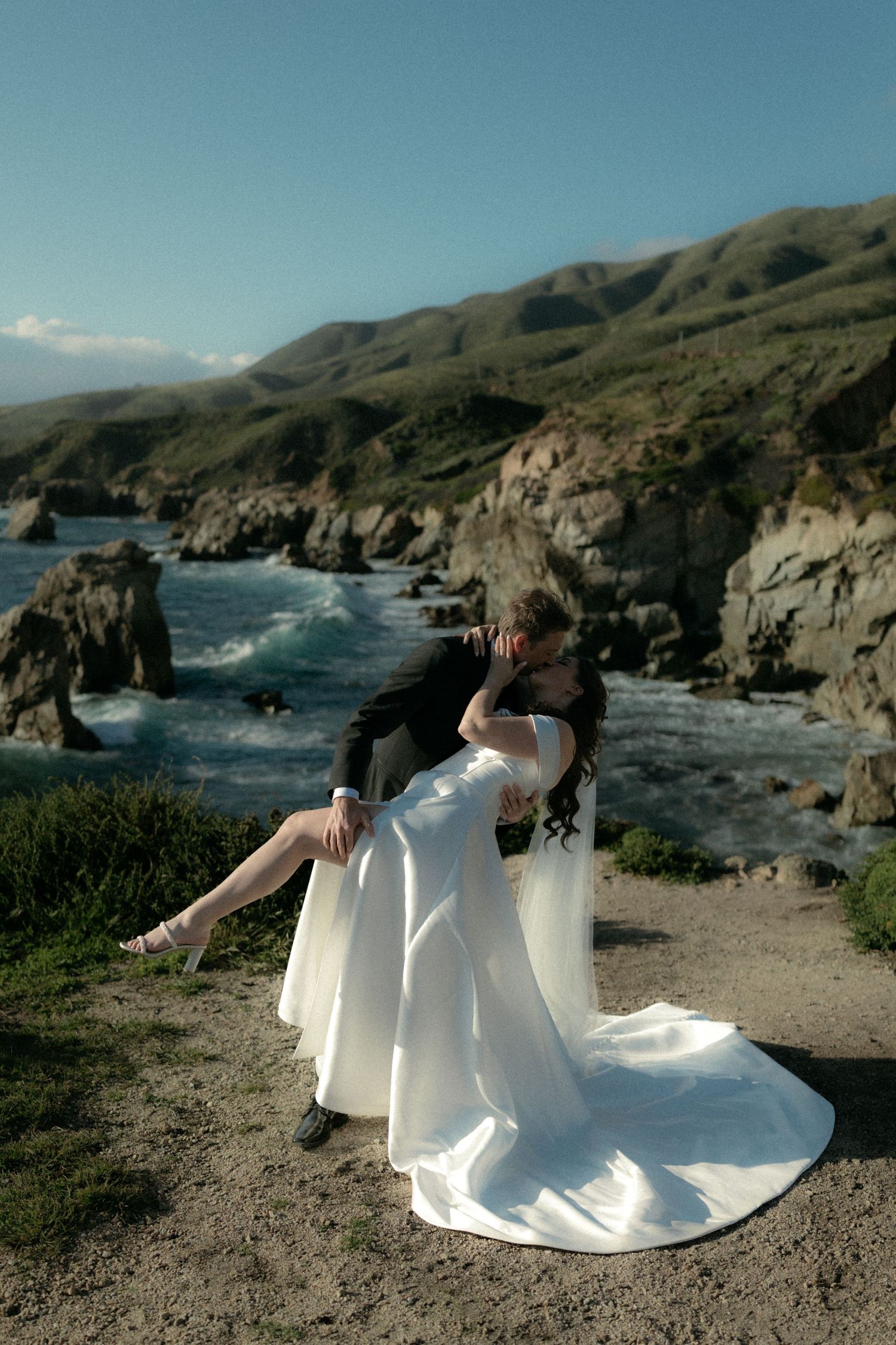 Bride and groom share a romantic embrace on a coastal cliff with ocean waves and rocky shoreline in Big Sur, California.