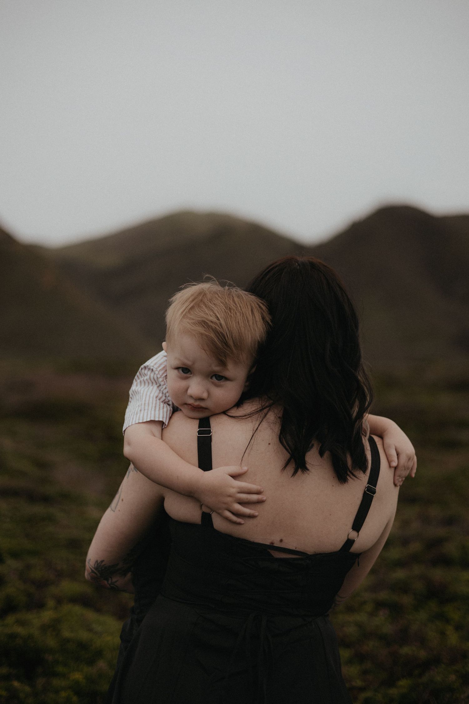 A mother with dark hair holds her blonde toddler in a green hilly landscape, the child looking at the camera.