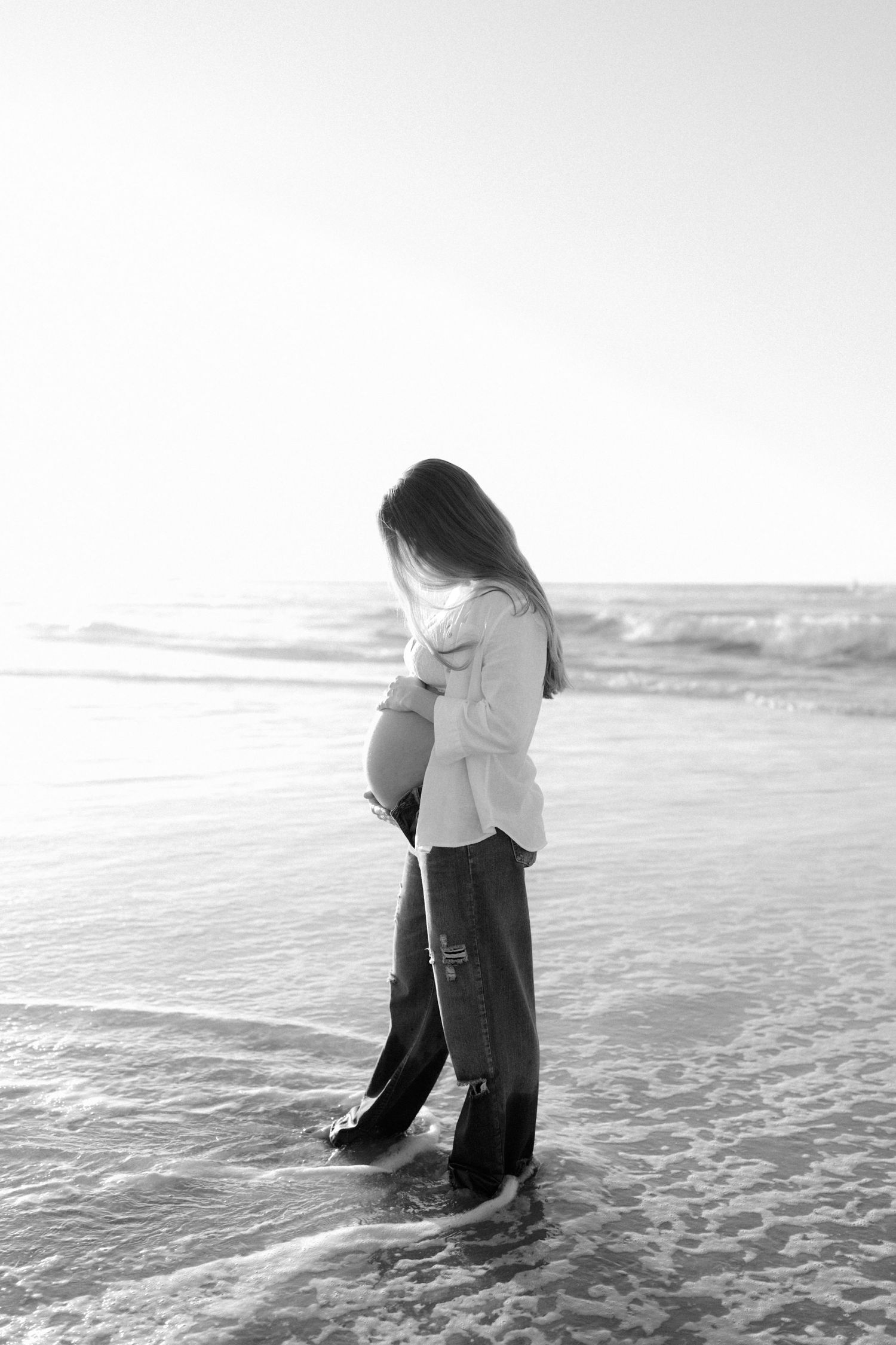 Black and white portrait of a pregnant woman standing alone on a beach, cradling her baby bump.
