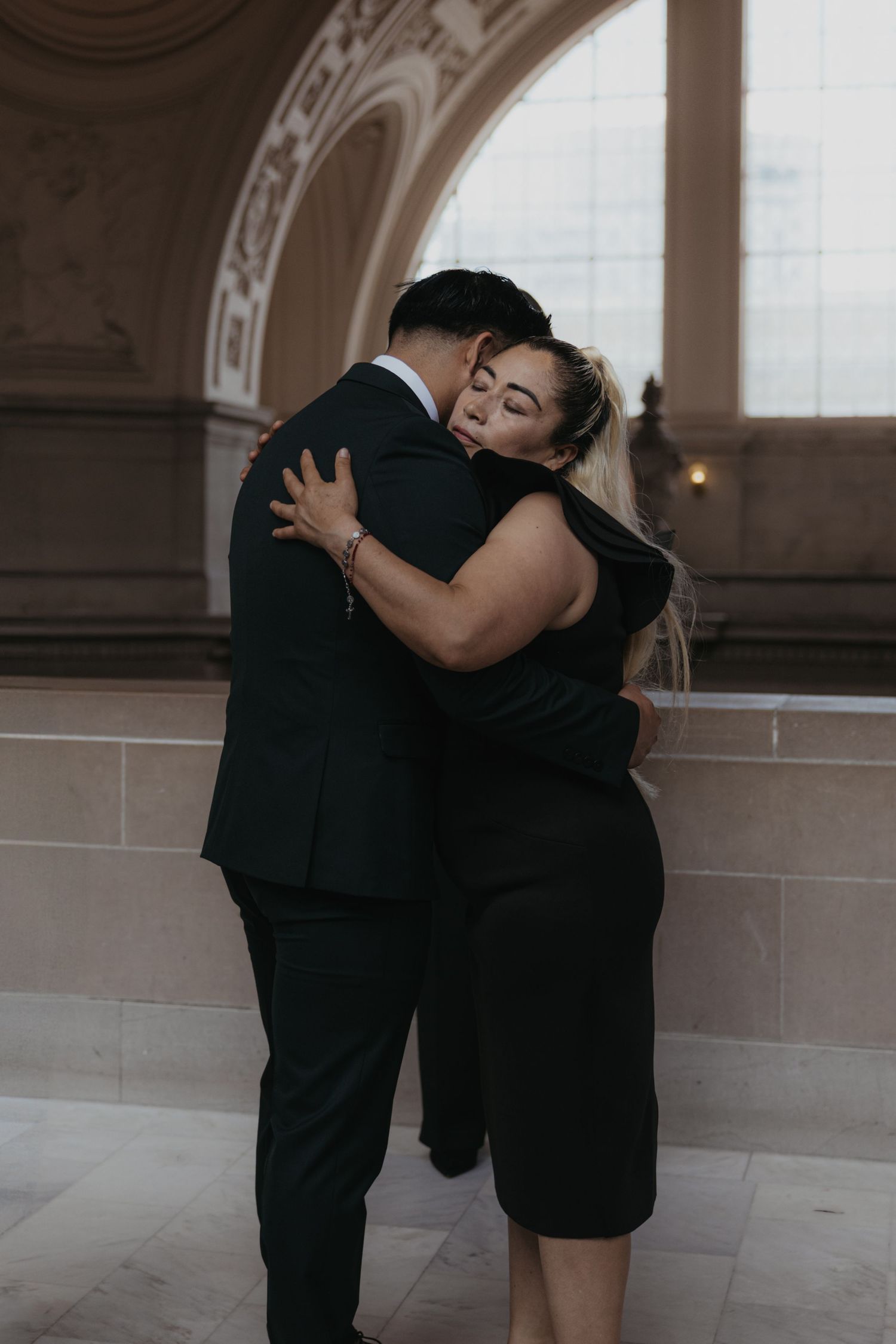 Two people embrace in formal attire against an elegant architectural backdrop.