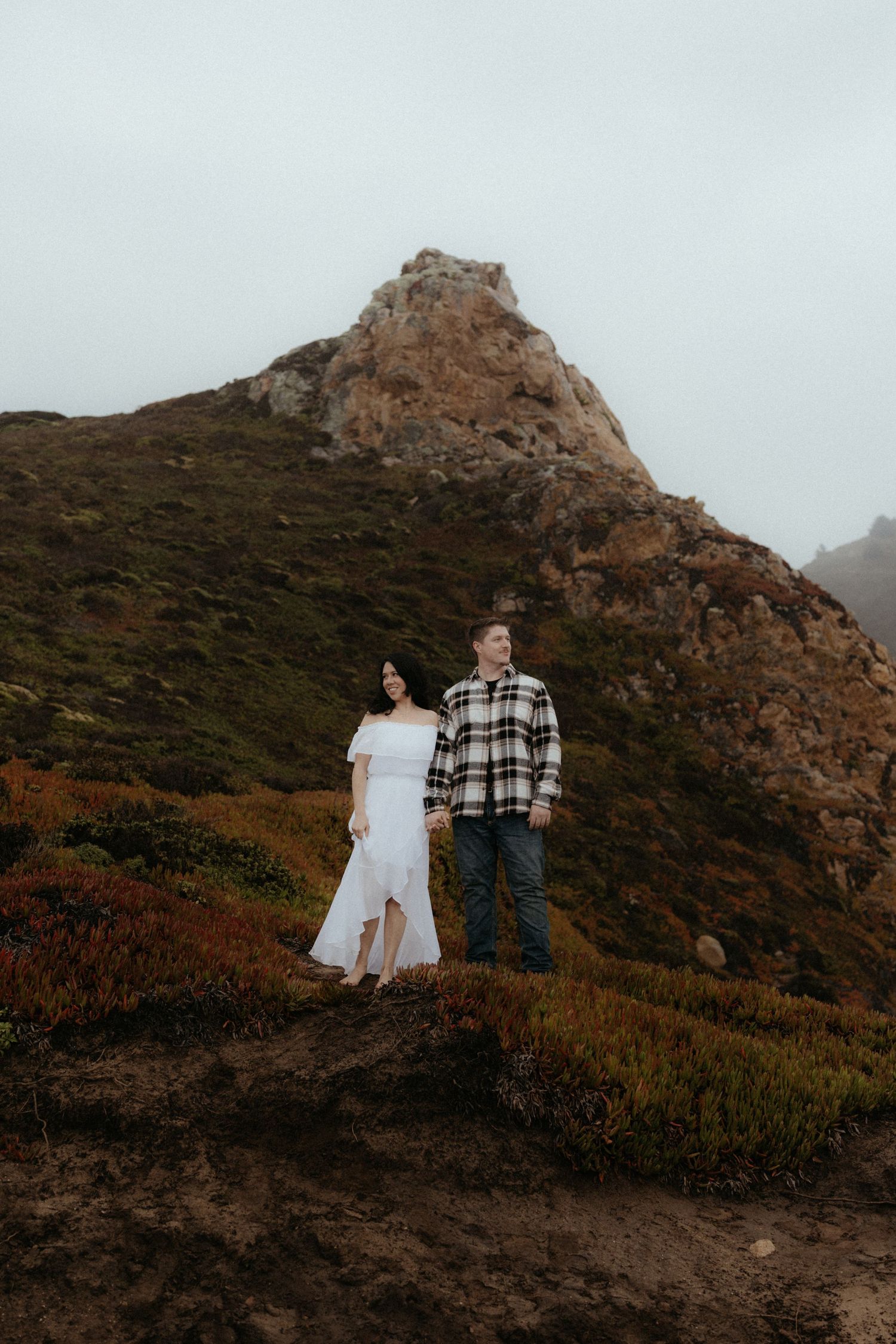 A couple poses together against a foggy mountain landscape with rugged terrain.