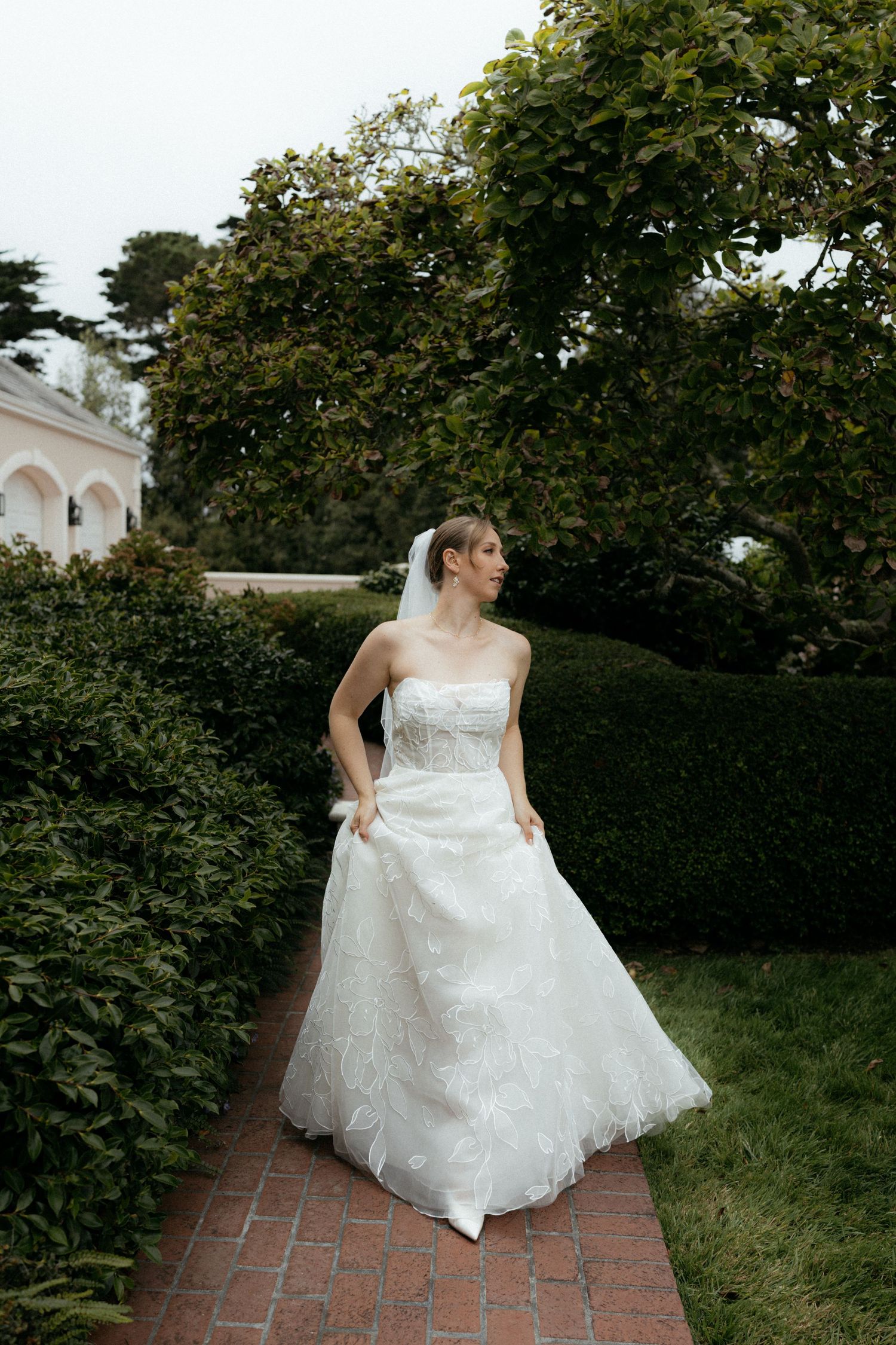 A bride in a strapless white lace ball gown and veil walks through a lush garden pathway.