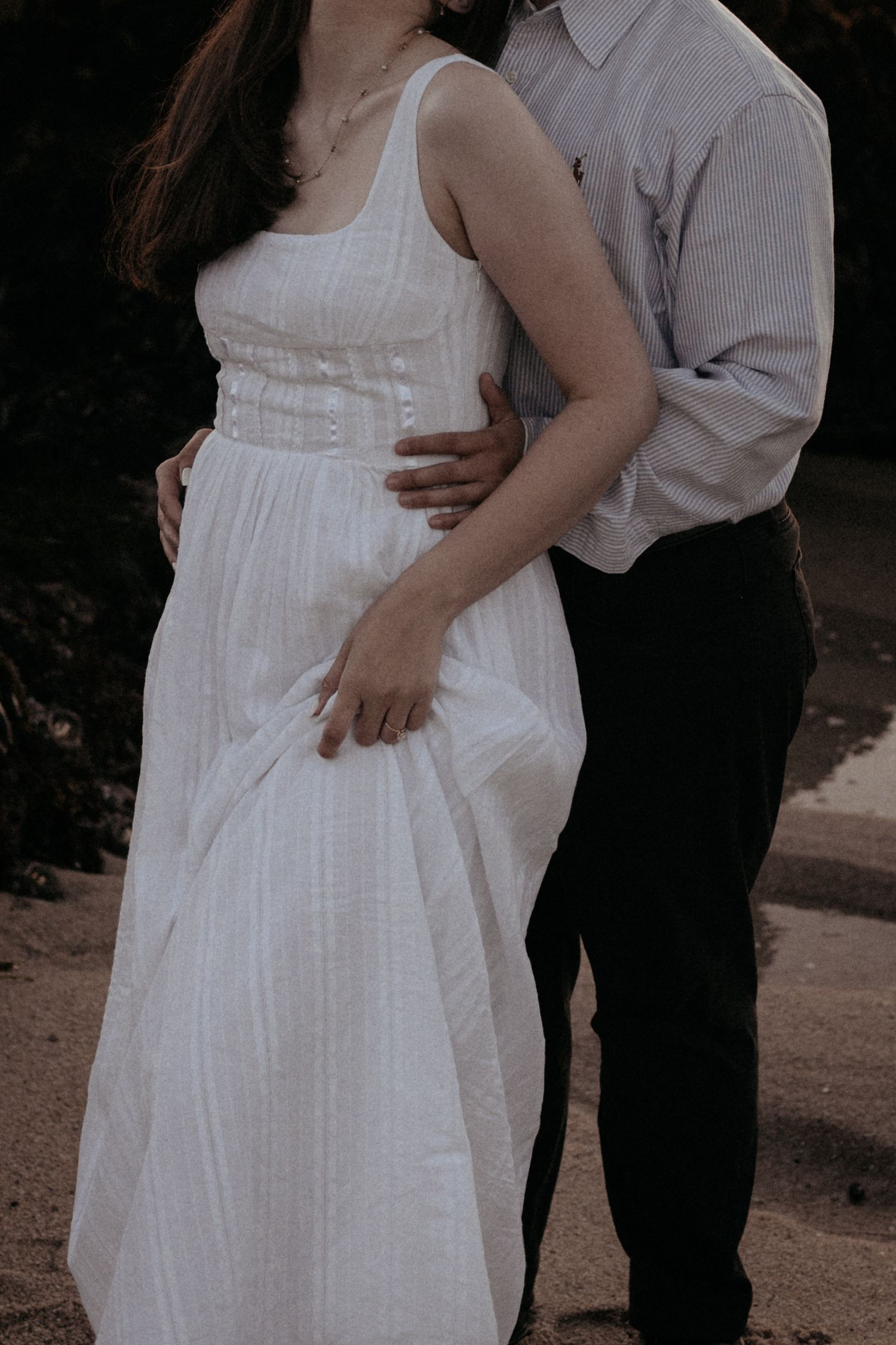 A couple embraces outdoors, woman in flowing white dress held by man in striped shirt and dark pants.