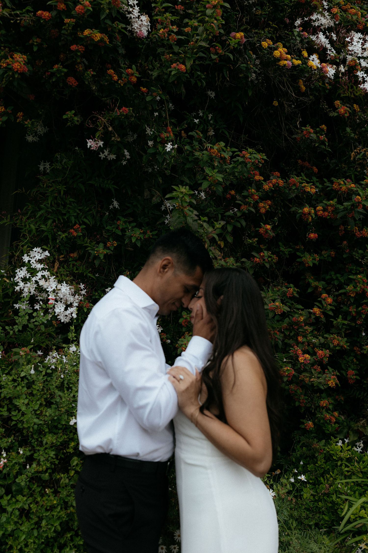 Romantic couple holding hands and touching foreheads in front of vibrant flowering garden wall.