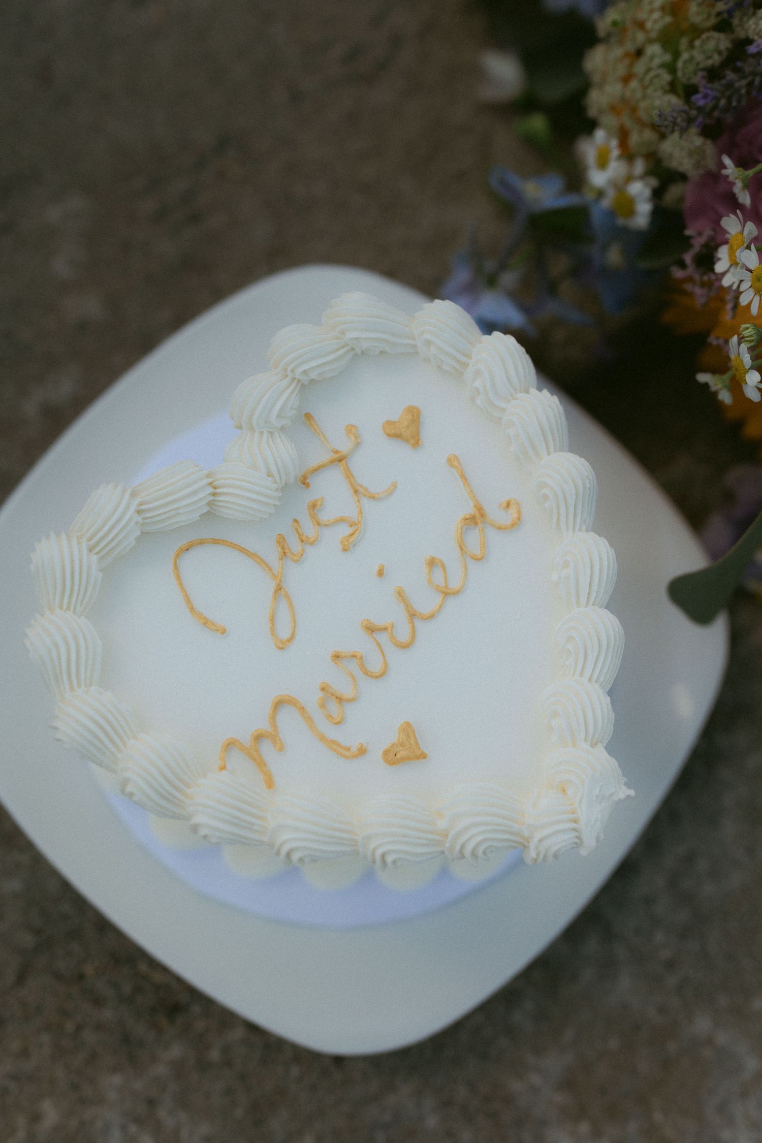 Heart-shaped white wedding cake with gold Just Married inscription beside wildflower bouquet.