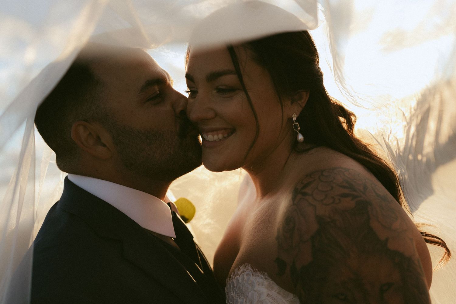 A groom in a navy suit kisses his tattooed bride on the cheek under a flowing veil at golden hour.