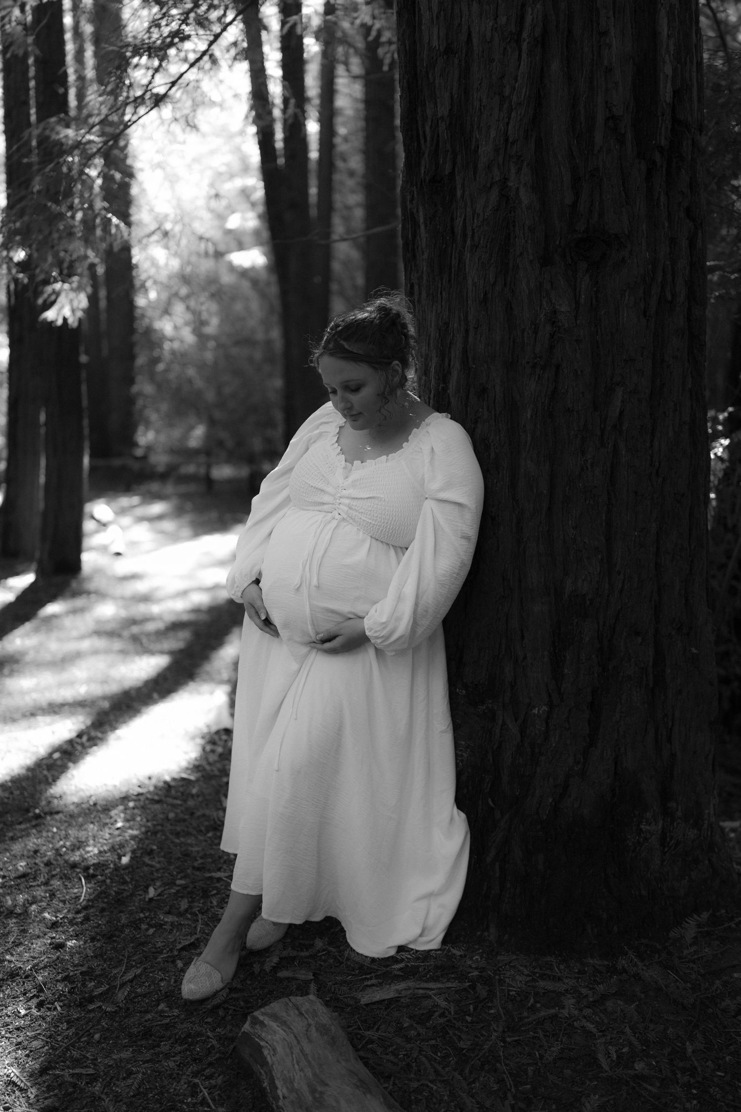 Person in flowing white dress sitting thoughtfully against a redwood tree in dappled forest light.