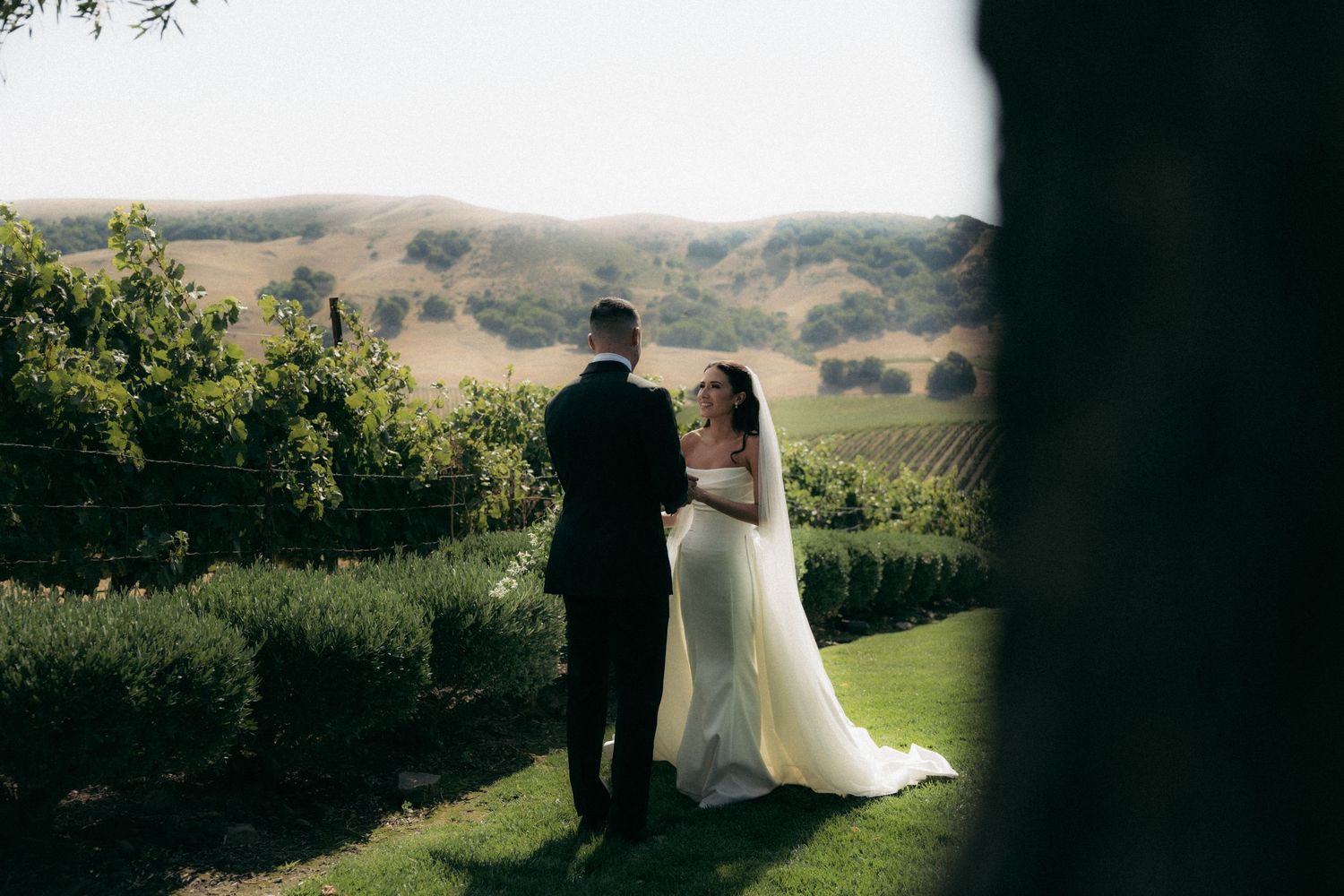 A couple stands together on a lush green hillside during their outdoor wedding ceremony at sunset.