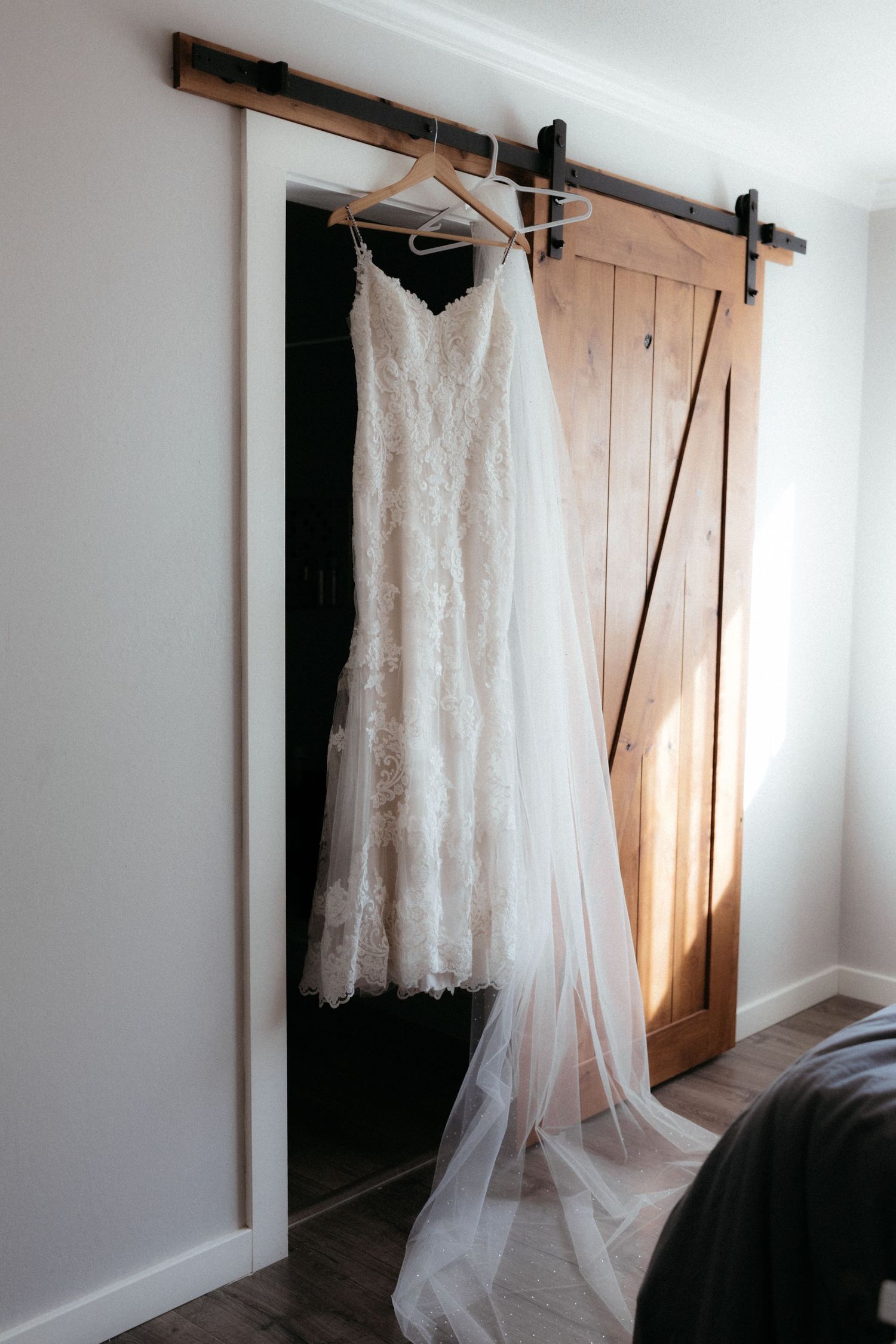 A white lace wedding dress hangs from a rustic barn door in a bright room with natural light streaming in.