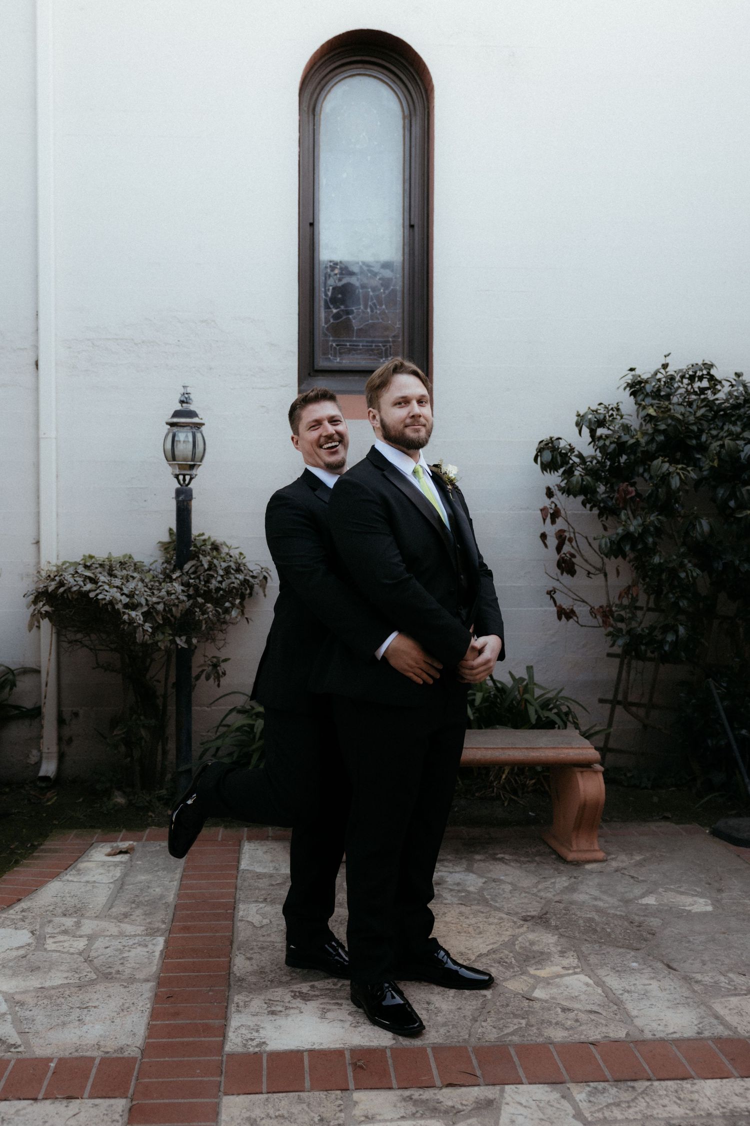 Two people in formal black attire pose playfully on a stone patio near white walls and garden shrubs.