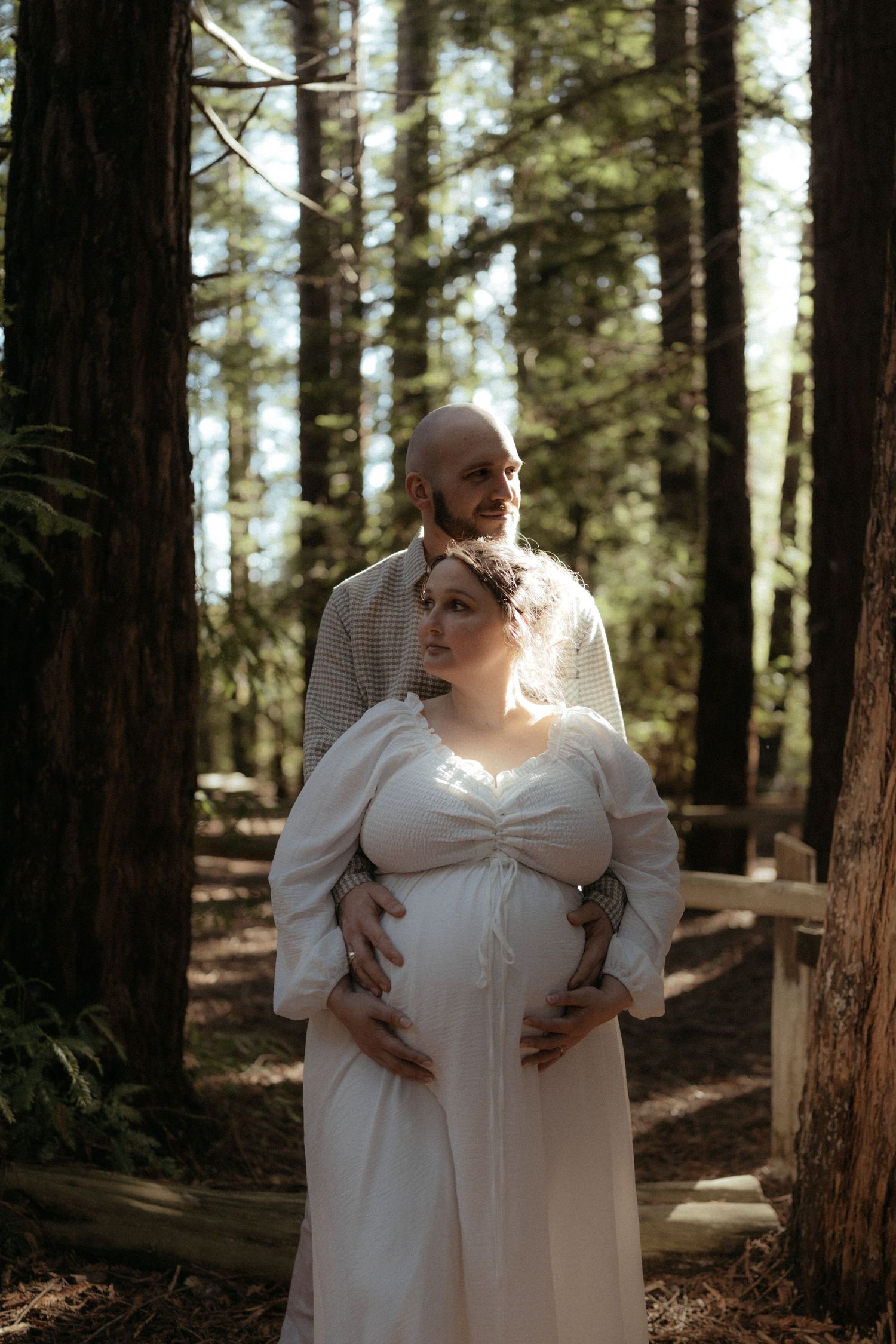 A couple embraces tenderly in a sunlit forest while wearing matching light-colored outfits.