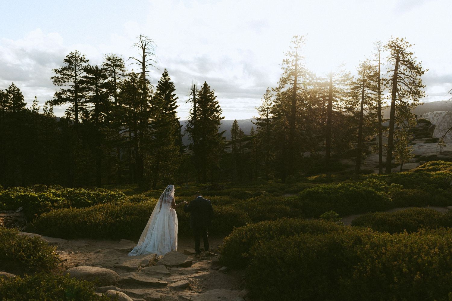 Bride and groom walking through manicured garden at sunset with pine trees and mountain views.