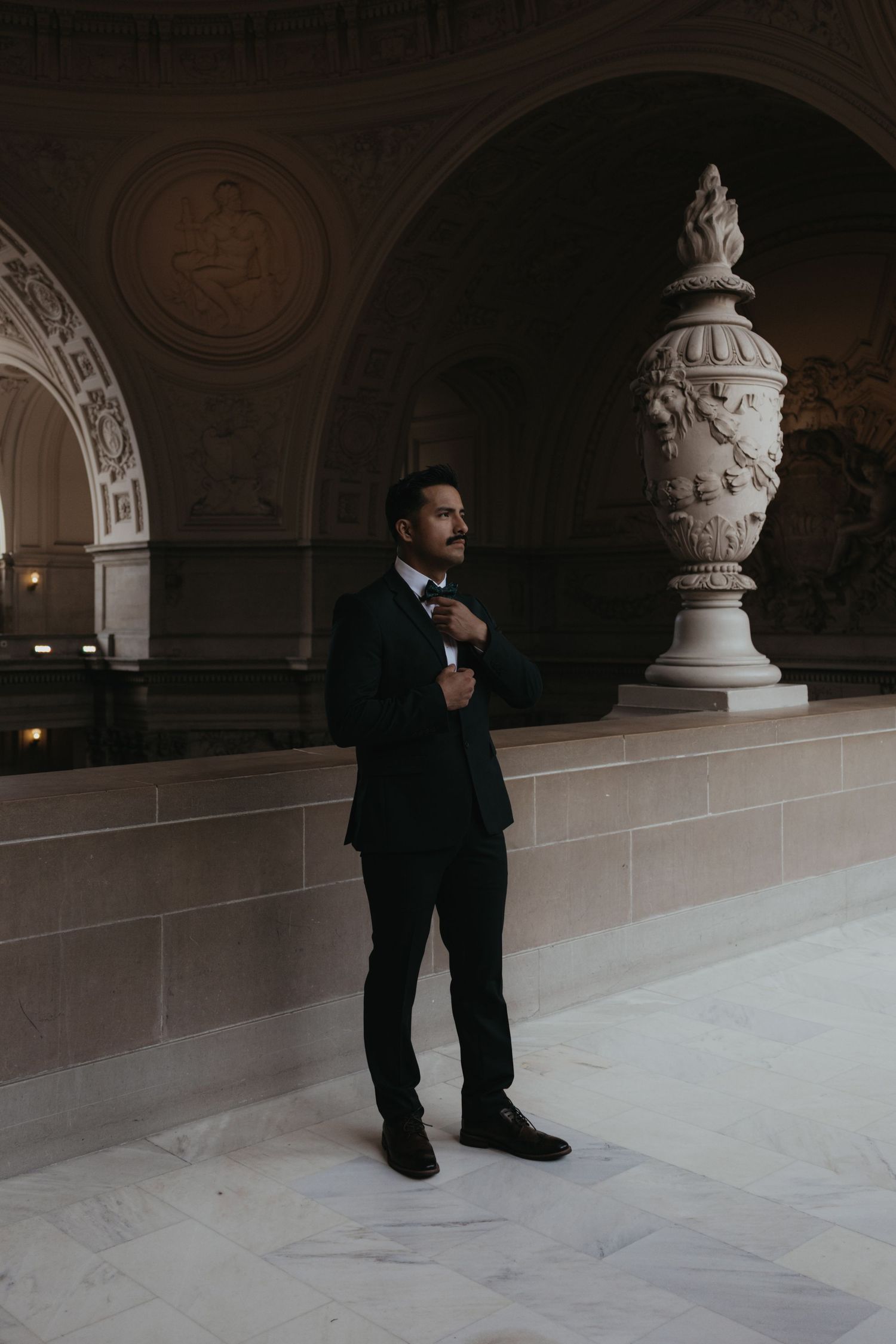 A groom in a black tuxedo adjusts his tie in an ornate marble hallway.