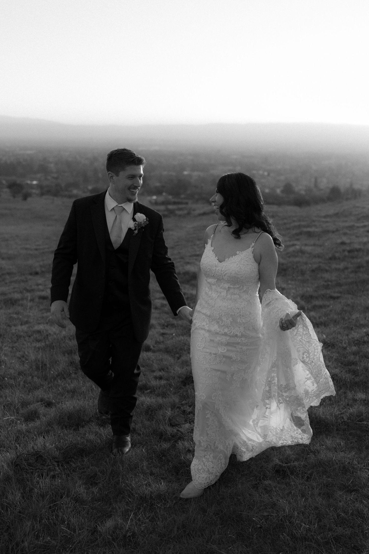 Black and white photo of newlyweds walking together across a hilltop meadow.