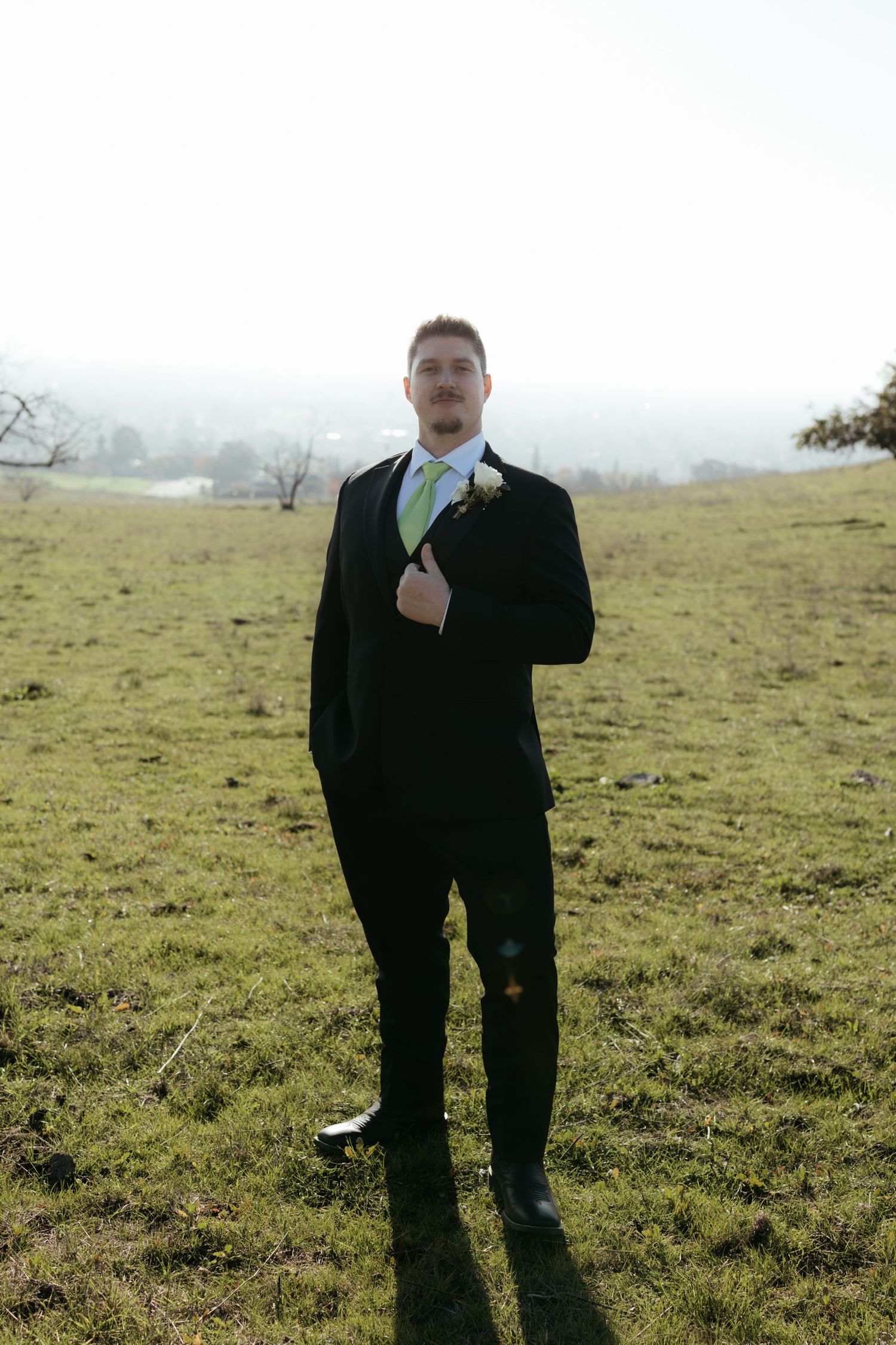 A groom in a black suit with lime green tie stands in a grassy field on a misty day.