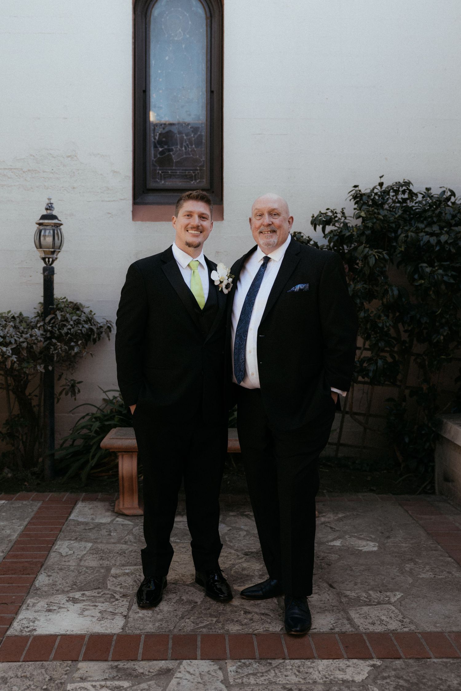 Two people in formal black attire stand together at a wedding venue with white walls and outdoor lighting.