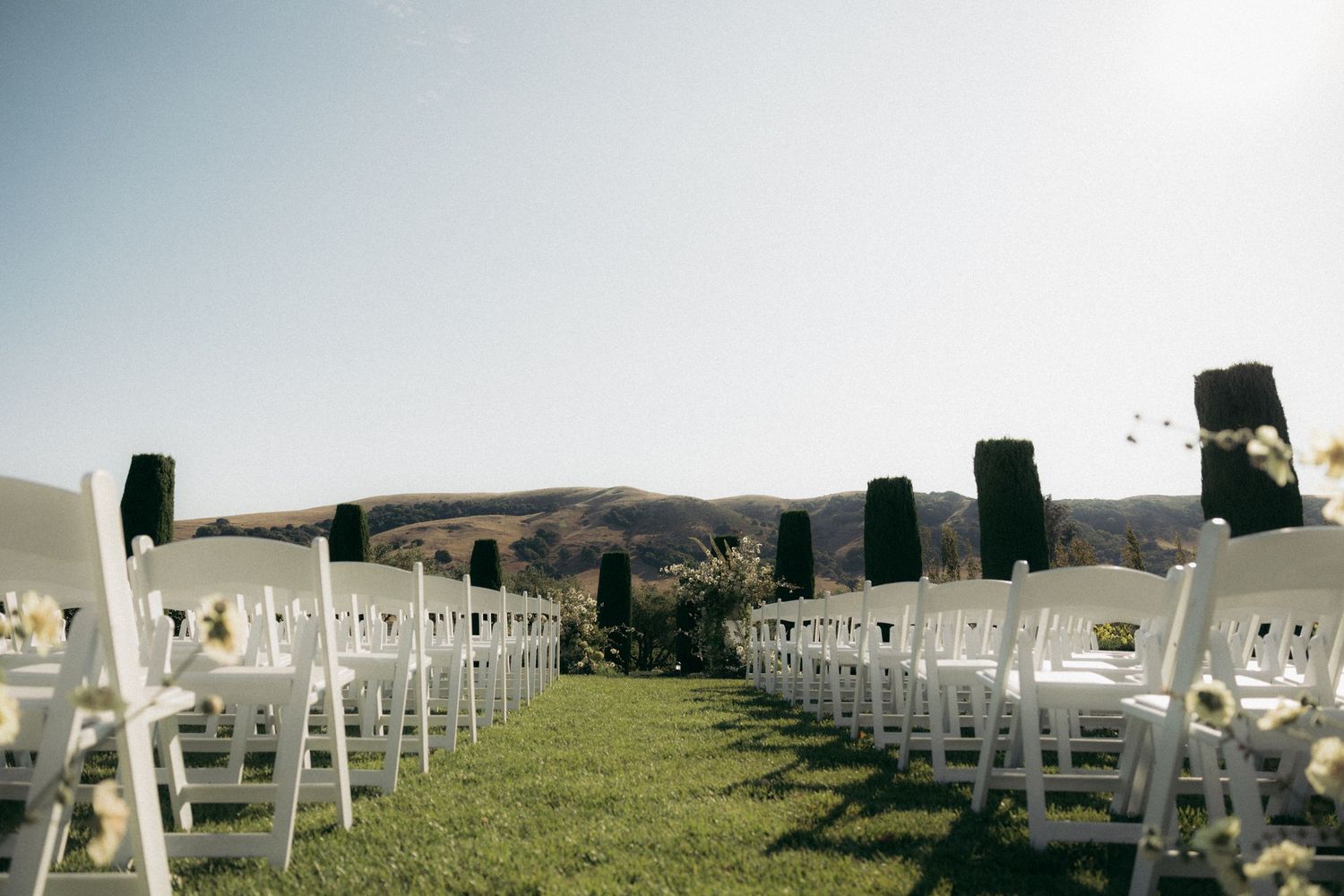 White wedding chairs arranged in rows on a grassy hillside with mountains in the background for an outdoor ceremony.