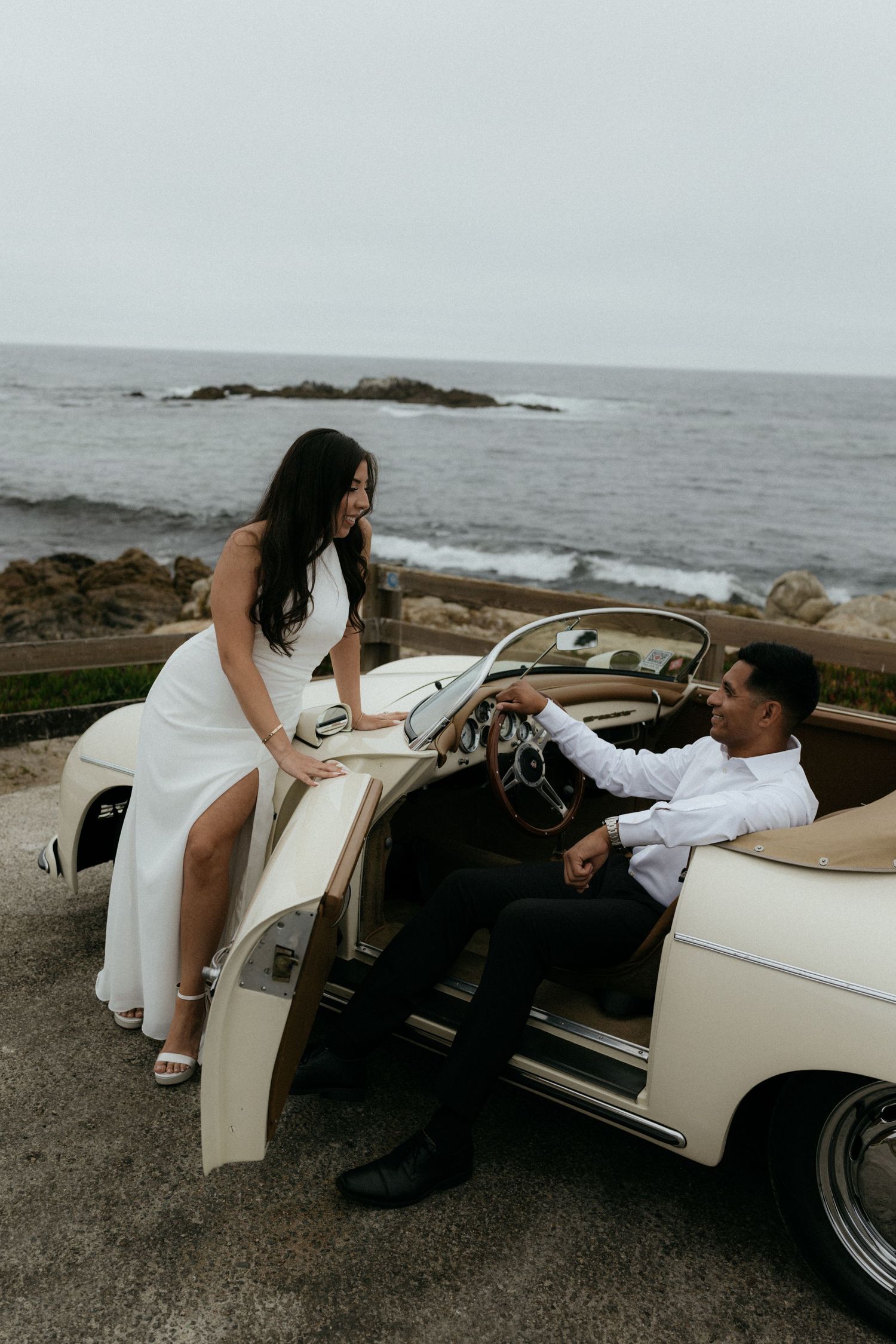 Couple in wedding attire posing with a vintage cream convertible car by the ocean.