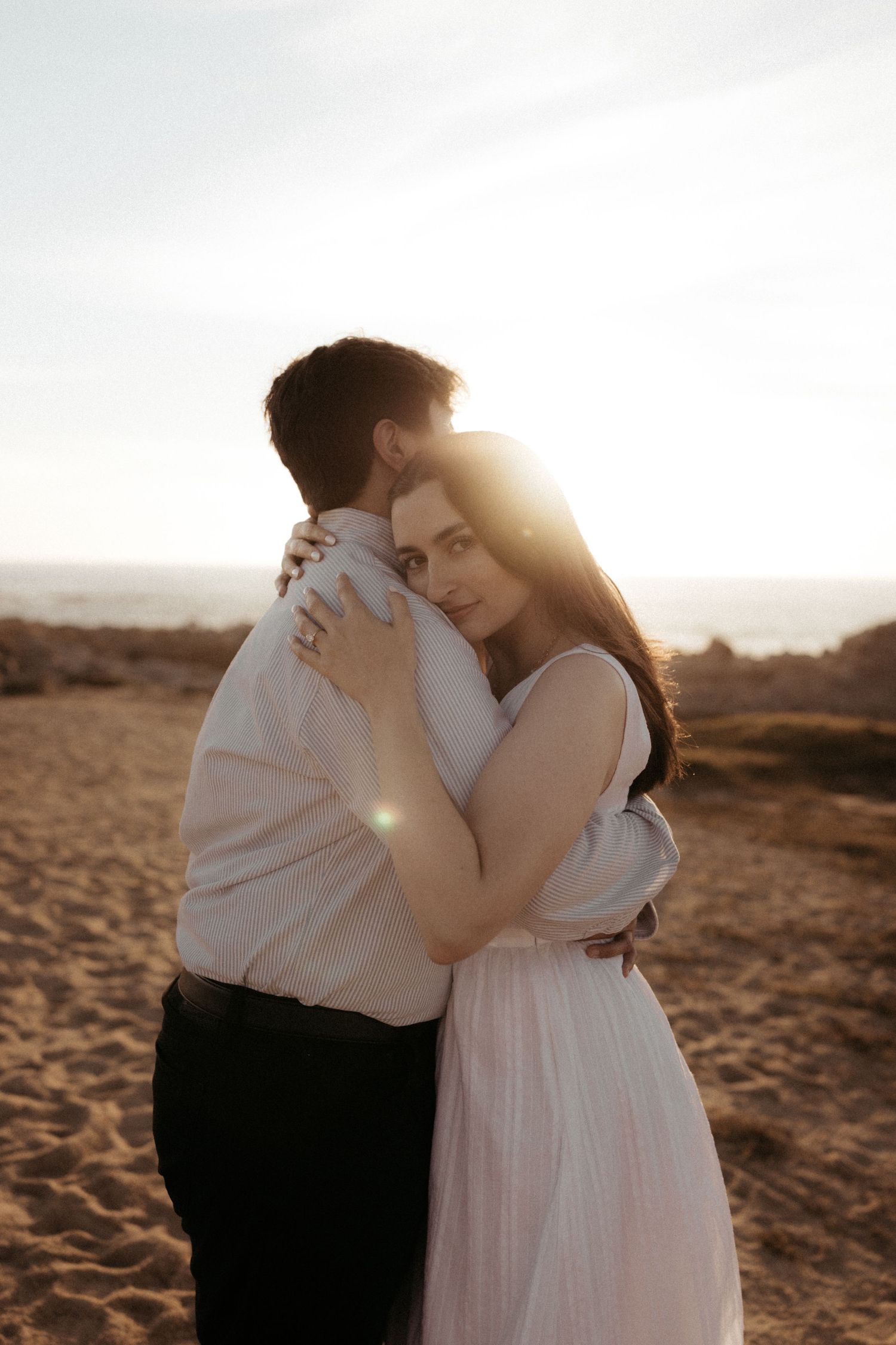 Couple embracing at sunset on sandy beach, woman in white dress gazing at camera with golden backlight.