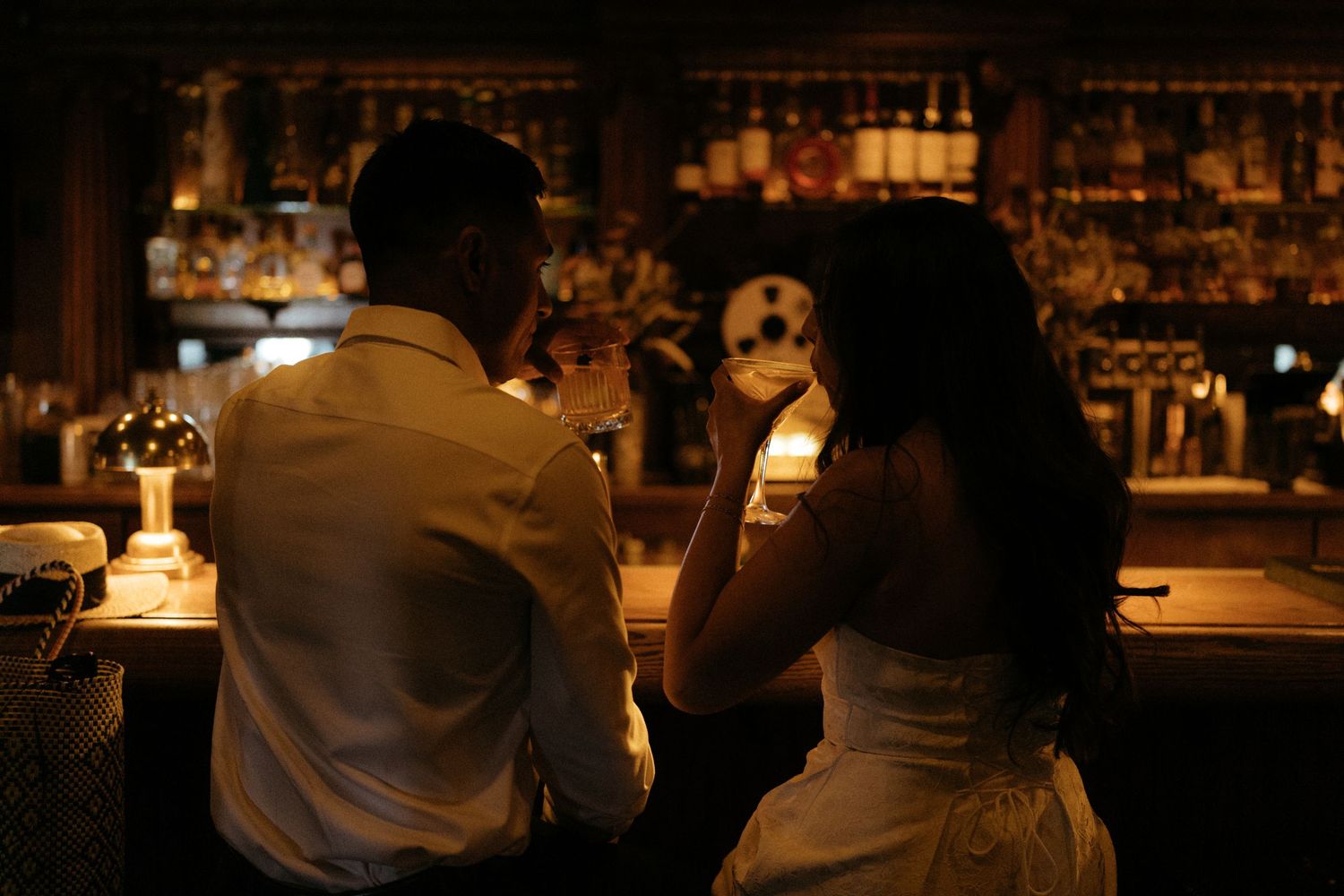 Couple sitting at a dimly lit bar, sharing drinks in a romantic atmosphere with bottles displayed in the background.