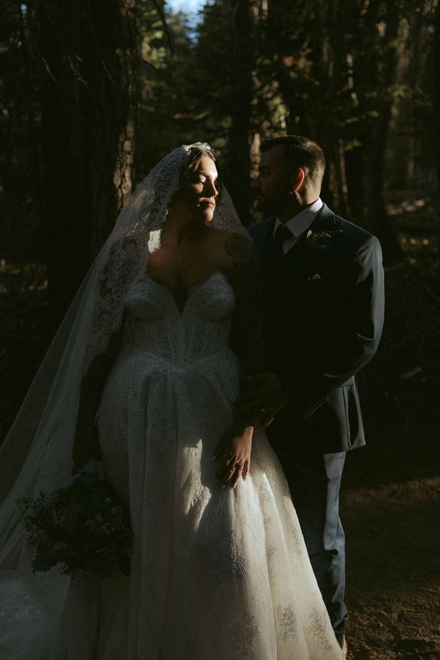 Bride in lace gown and veil holds bouquet beside suited groom in sunlit forest wedding portrait.