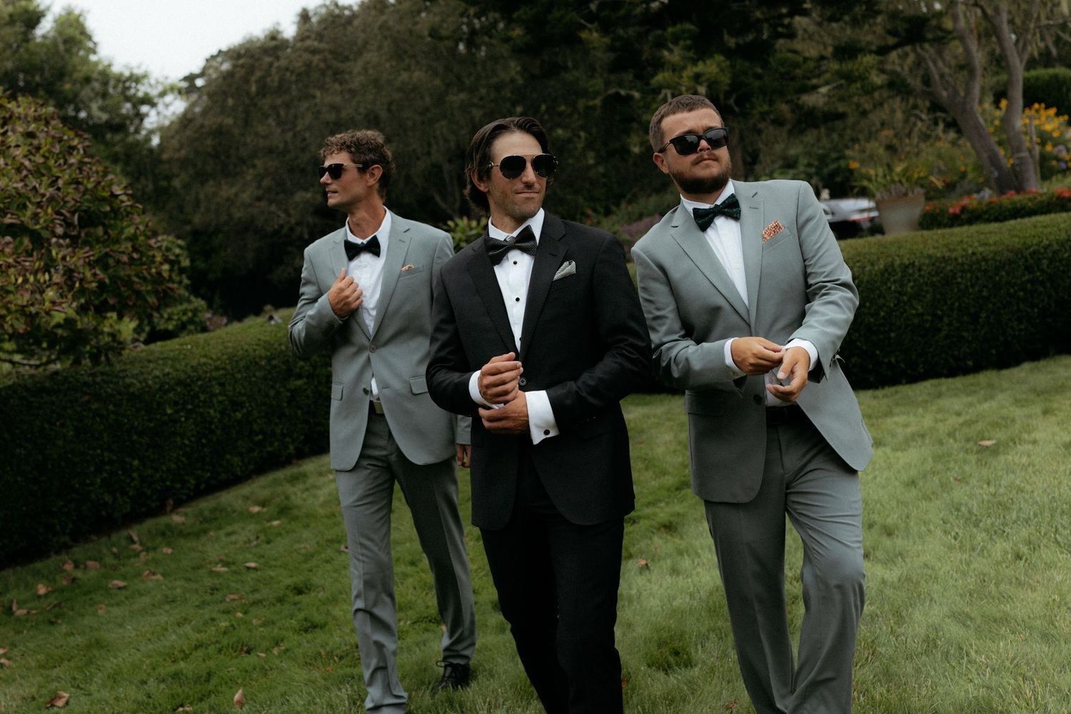 Three groomsmen in gray suits and bow ties wearing sunglasses walk confidently across a green lawn.