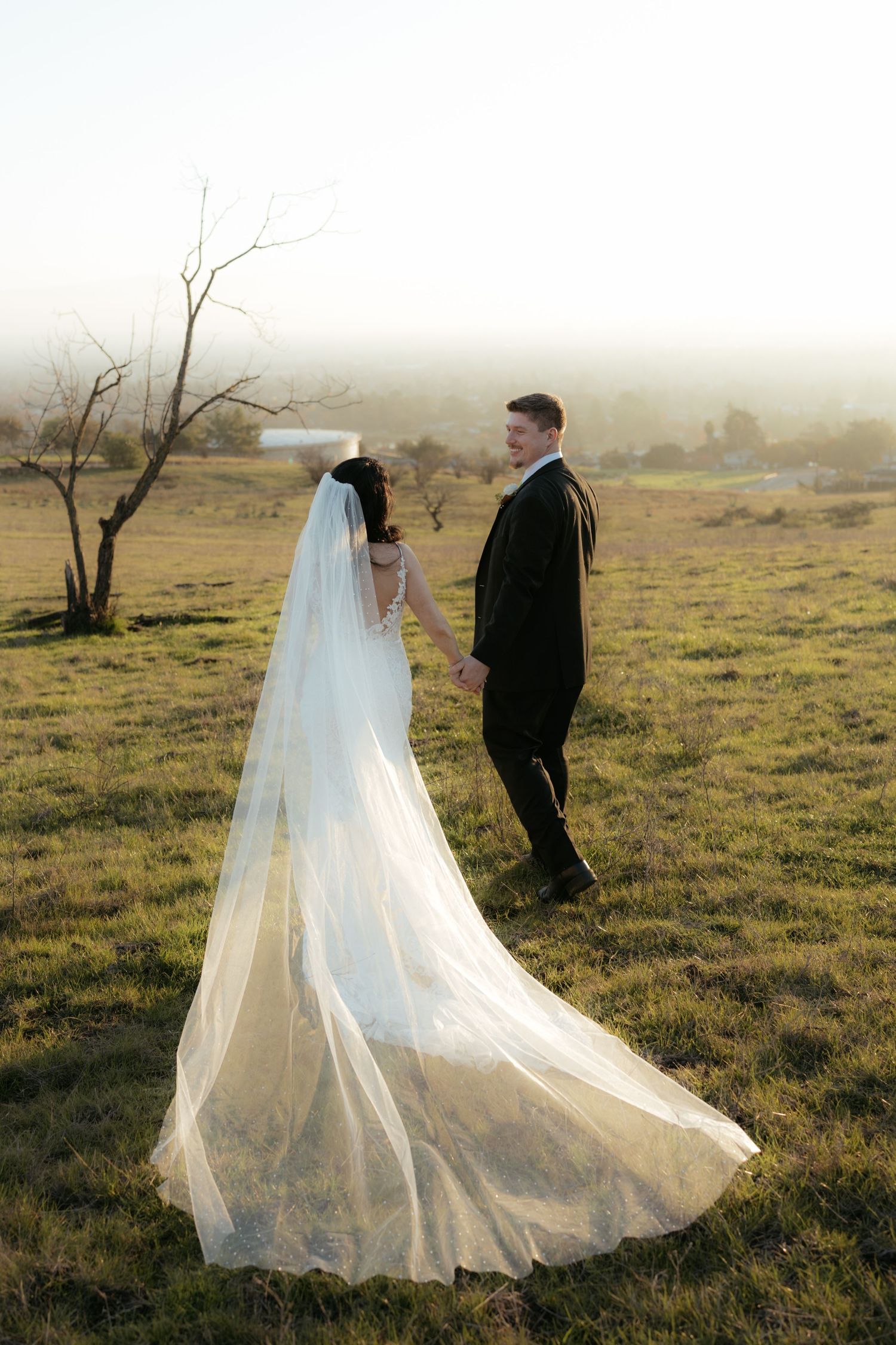 Wedding couple walking in a field at sunset with bride's flowing veil trailing behind.