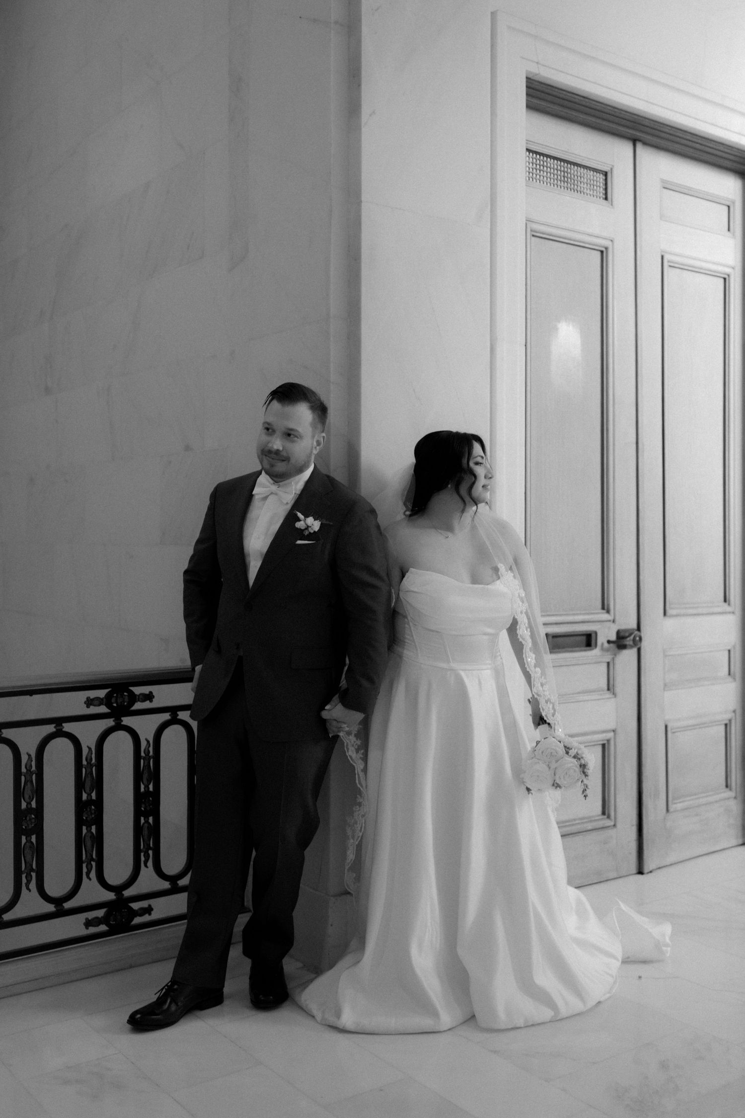 A black and white photo of a couple in wedding attire standing back to back in front of elegant white doors.