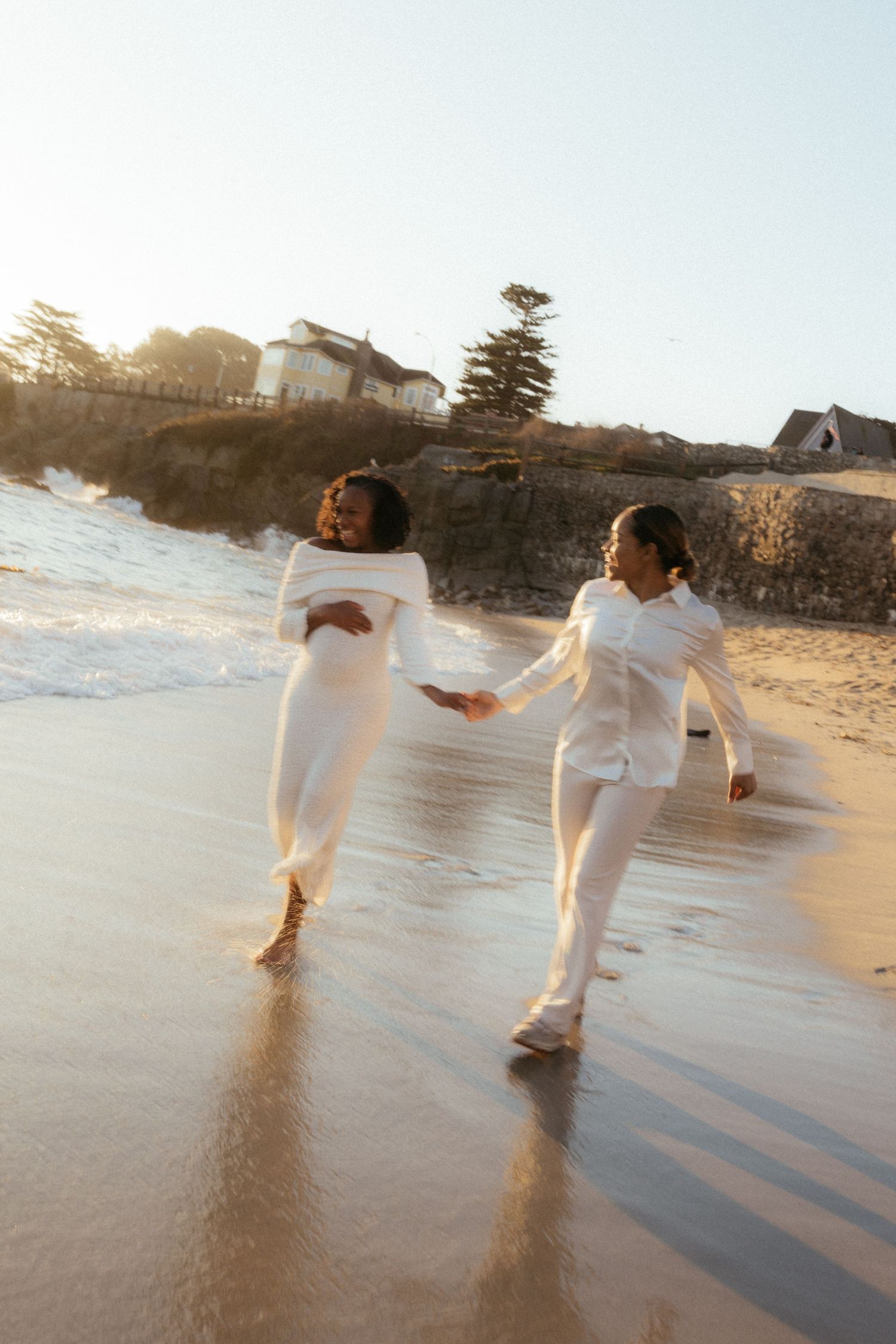 Two people in white outfits holding hands while walking along a beach at sunset with houses visible in background.