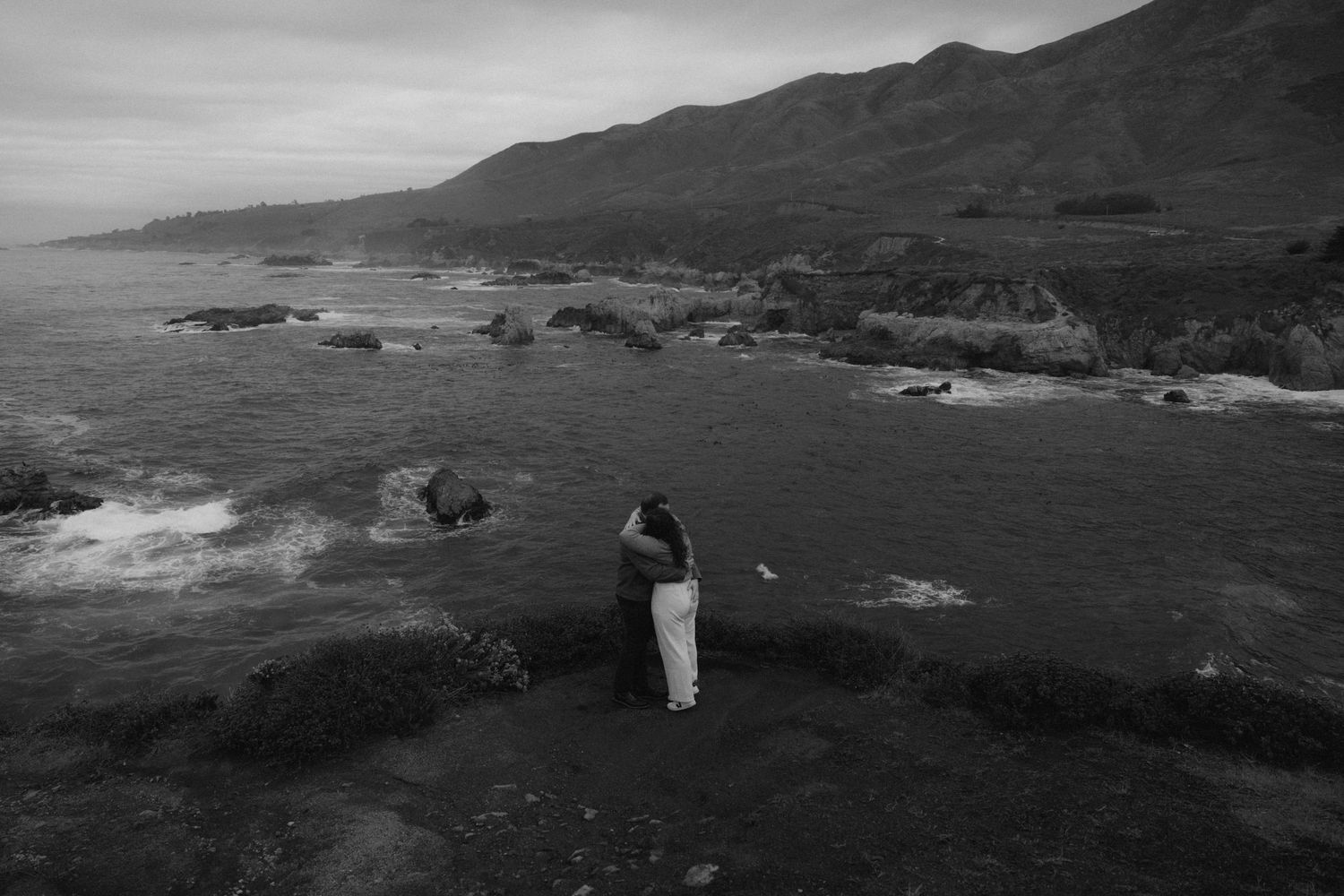Black and white panoramic coastal landscape showing rugged cliffs and rocky shoreline against a dramatic mountain backdrop.