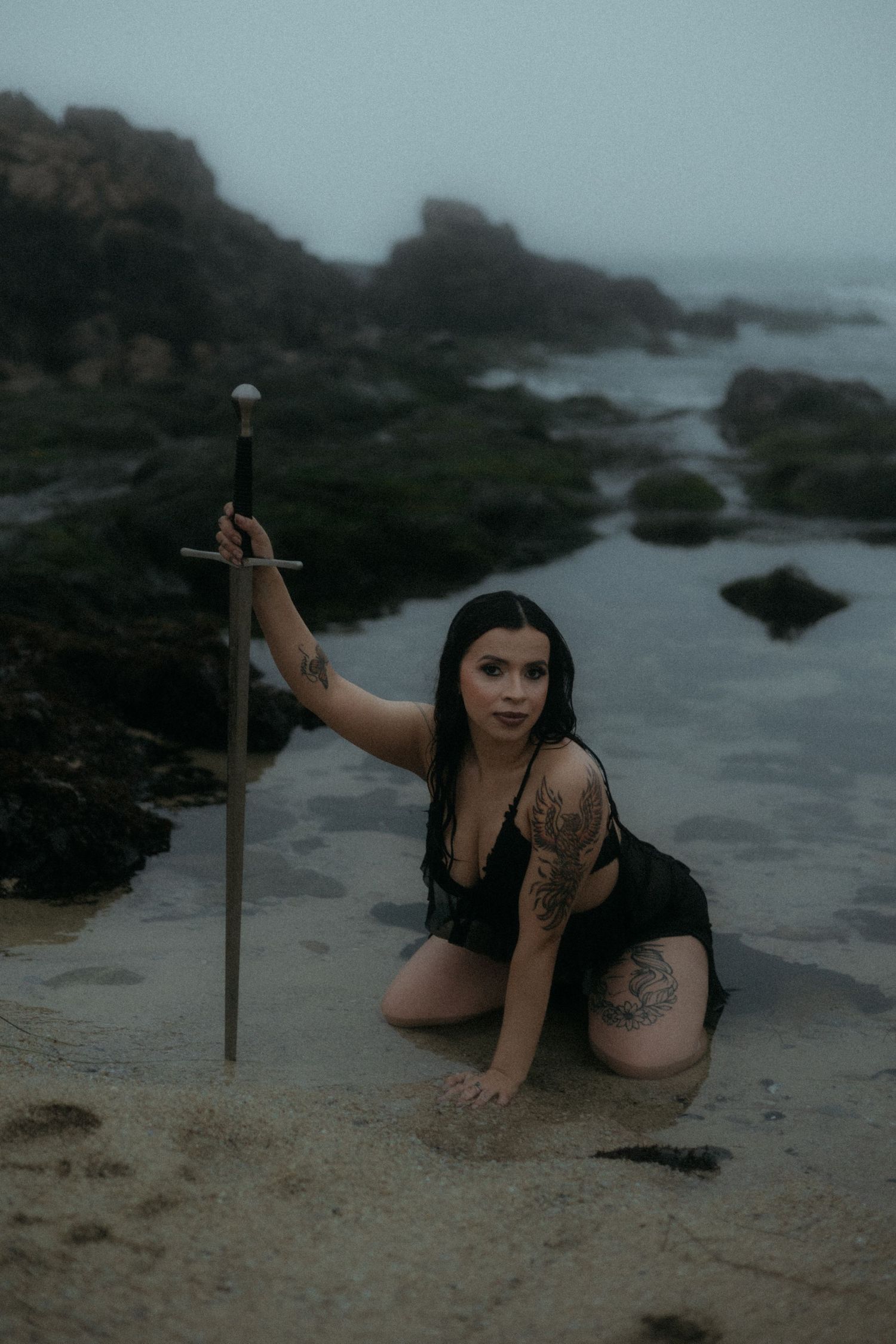 Dark-clad figure poses near wooden cross on misty beach with rocky outcrops.