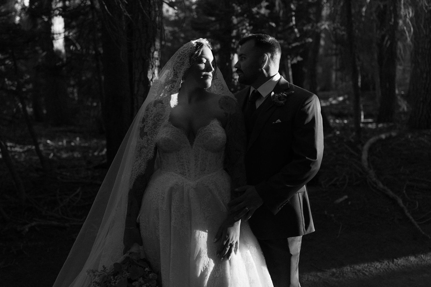 Black and white wedding photo of bride in flowing gown and groom in kilt standing together in a forest.
