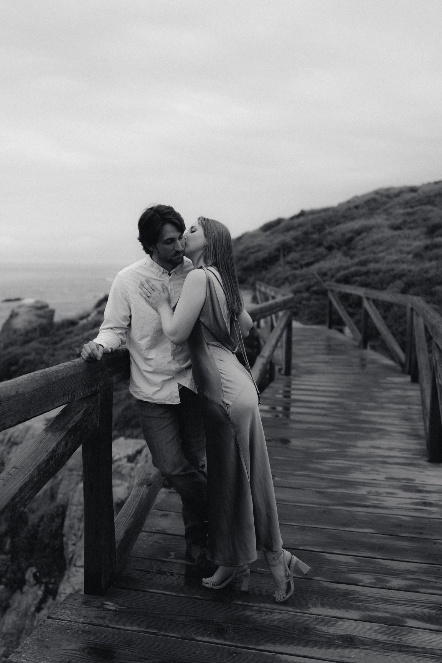 Black and white photo of a romantic couple kissing on a wooden boardwalk overlooking the ocean.