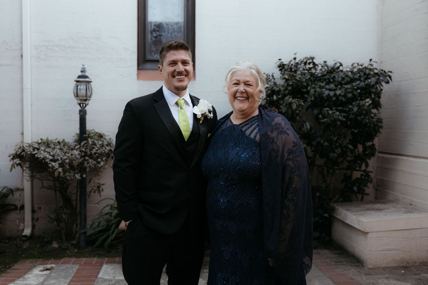 Two people in formal attire stand together smiling outside a building with a decorative lamp post and shrubs.