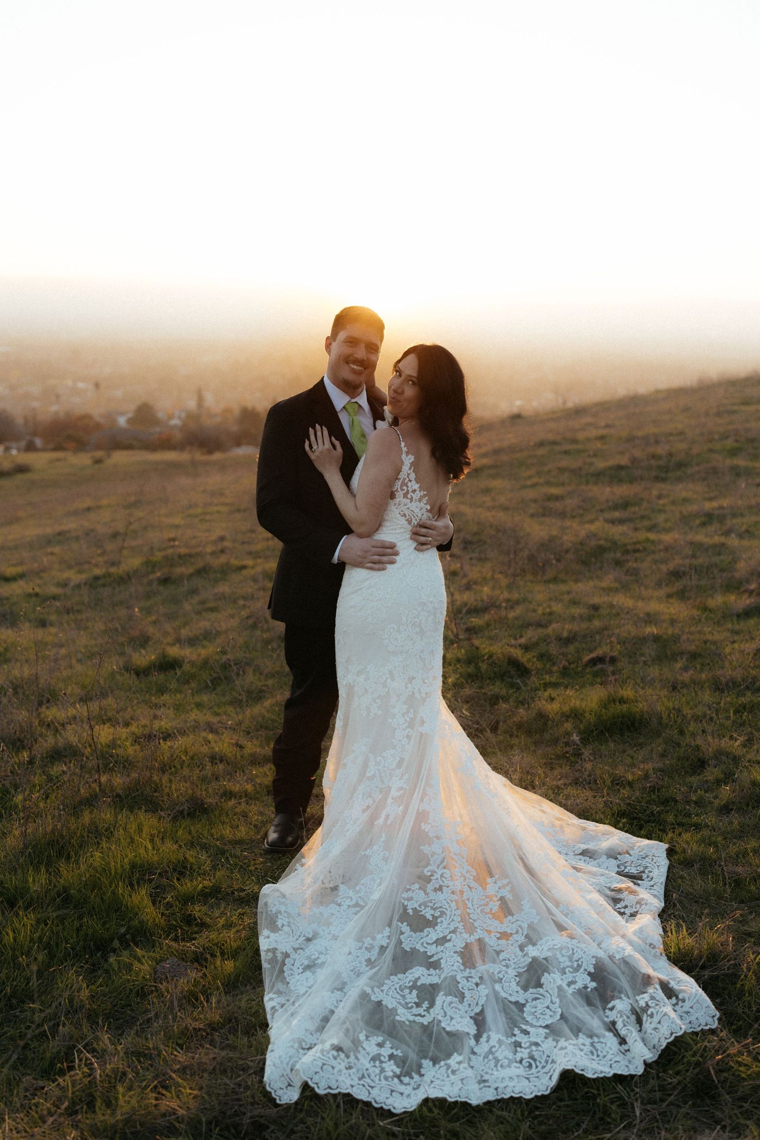 Wedding couple poses on hillside with bride's lace dress train beautifully displayed in sunset light.